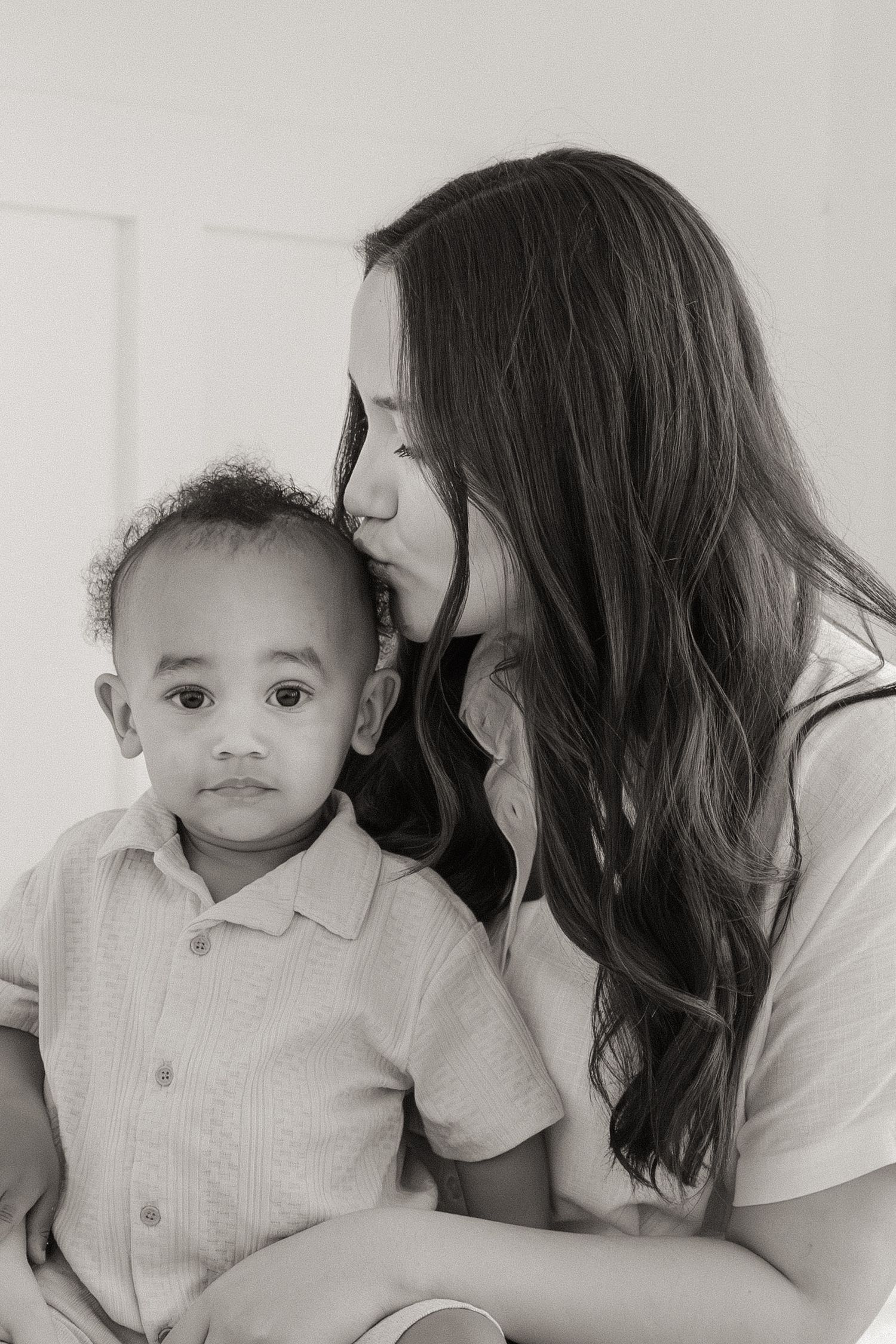 Black and white photo of a woman with long hair kissing a young toddler boy on the cheek.