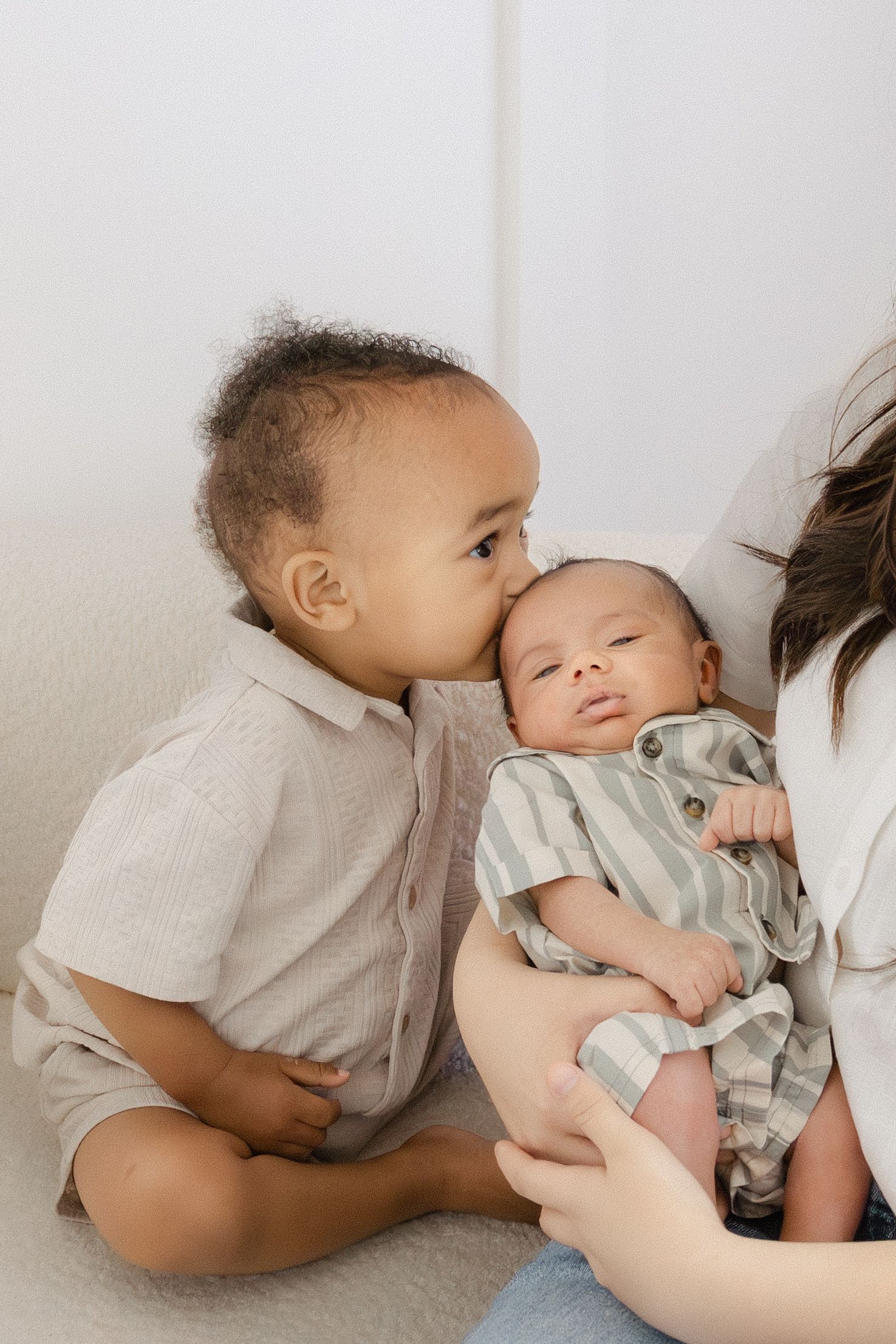 A toddler boy gently kisses a newborn baby sibling held by their mother on a soft white couch.