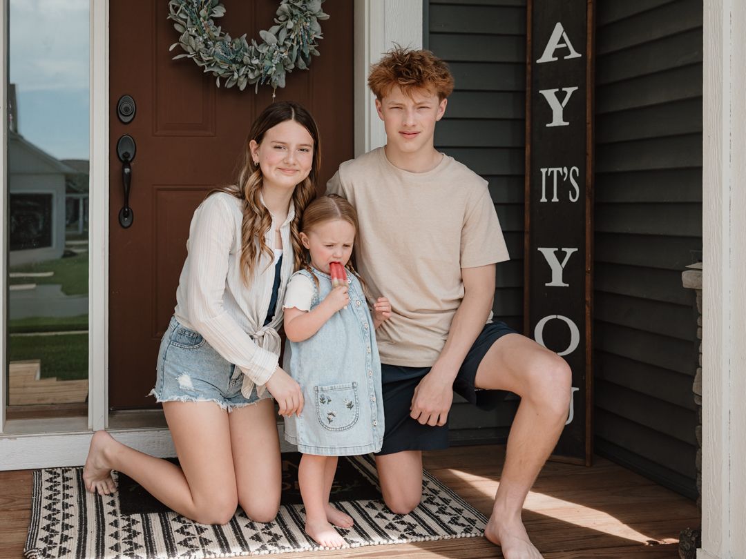 Two teenagers and a young girl in denim overalls posing together on a sunny home front porch.