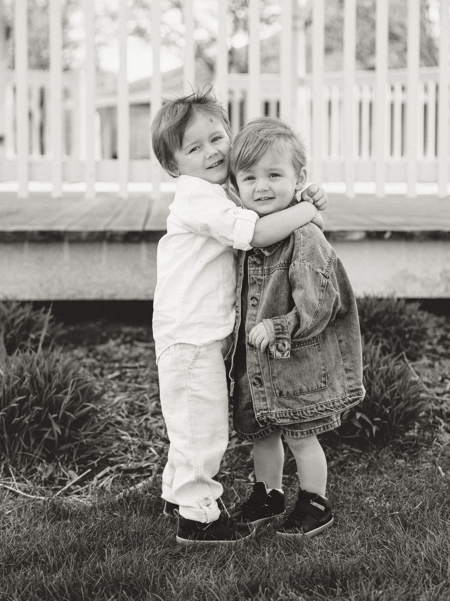 Two young children hugging outdoors in black and white photo, boy in white outfit embracing toddler in denim jacket on grass.