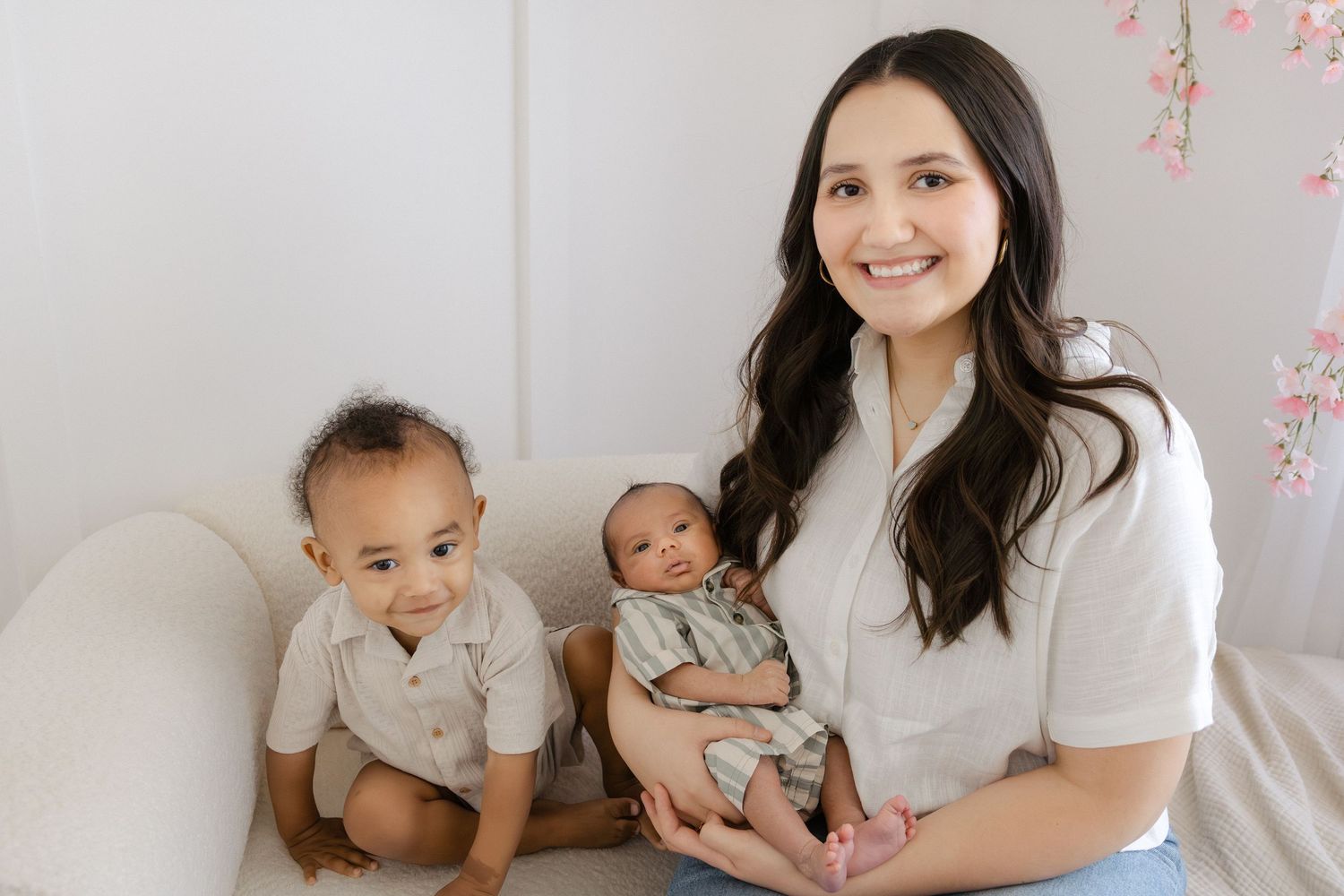 A smiling woman holds a newborn baby while a toddler sits beside her on a white couch in a bright studio.