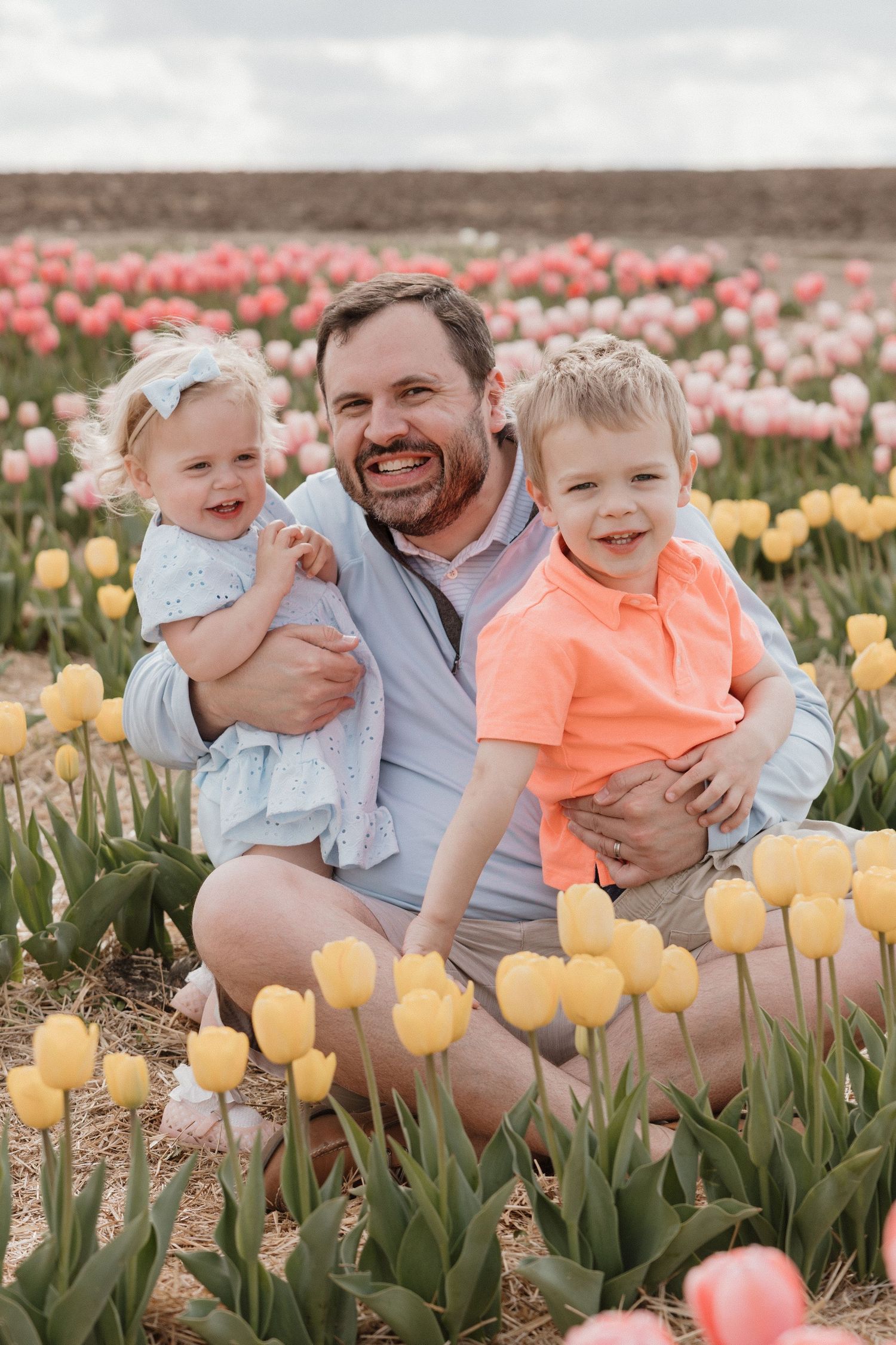 A smiling father sits in a colorful tulip field holding two young toddlers, a girl in blue and a boy in orange.
