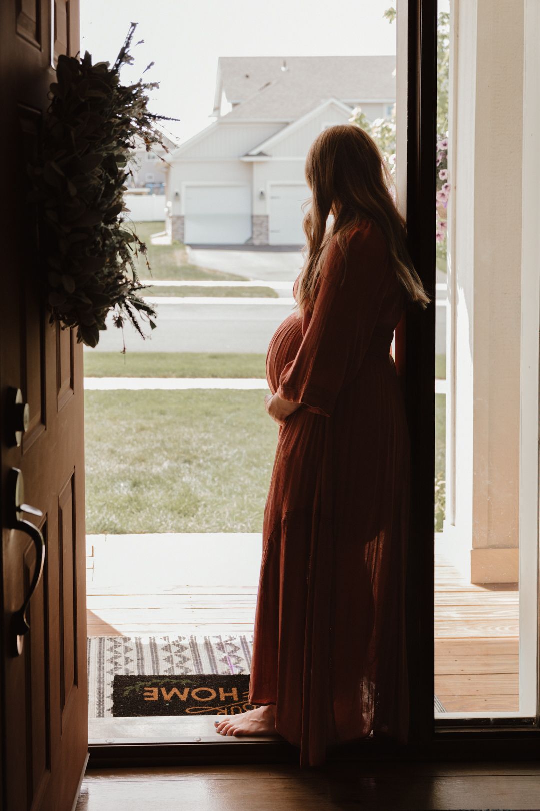 Pregnant woman in a rust-colored maxi dress standing in an open doorway, cradling her baby bump.