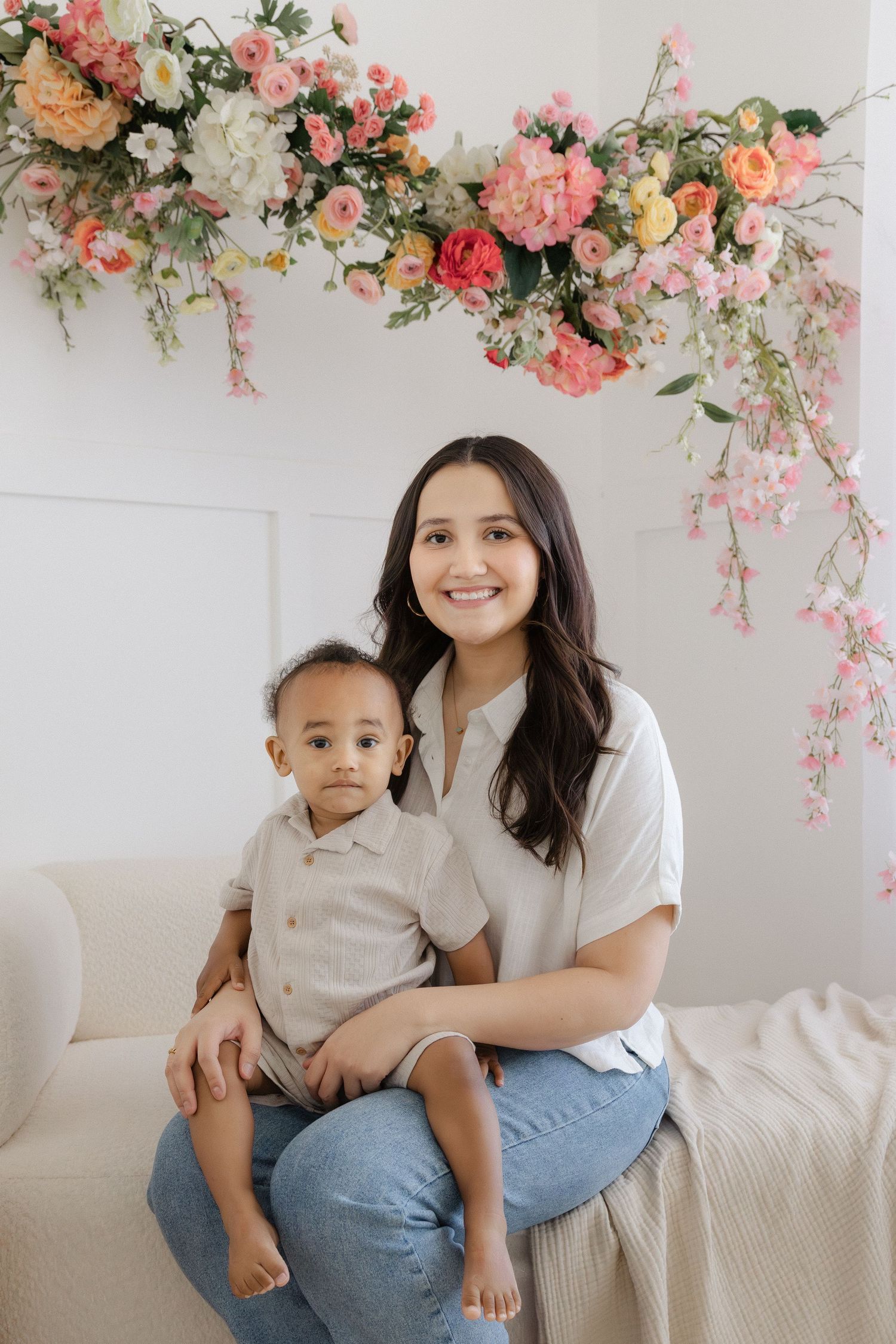 A smiling woman in a white shirt holds a toddler on a couch beneath a colorful floral arrangement.