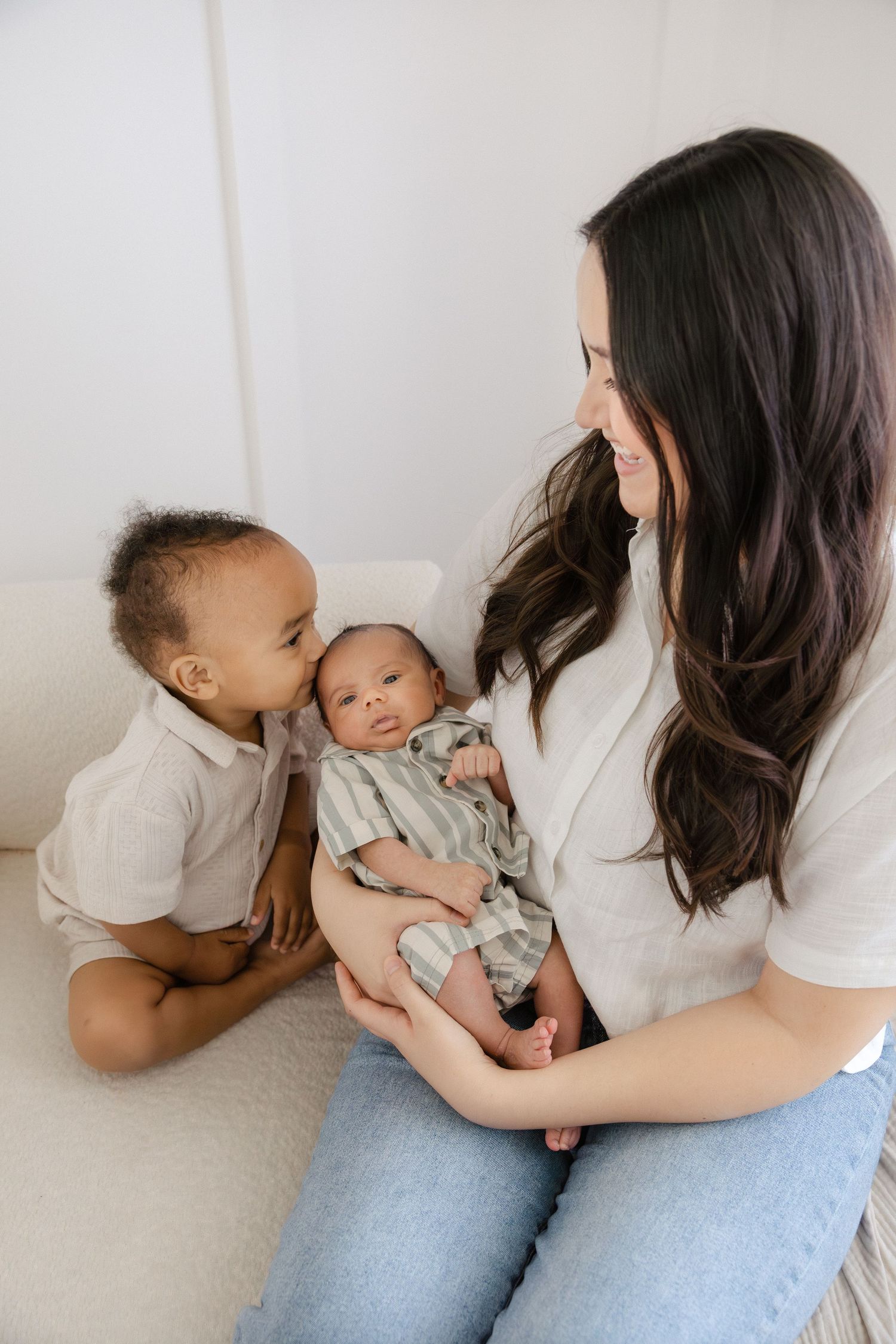 A mother with long dark hair holds her newborn baby while her toddler son leans in to give a gentle kiss.