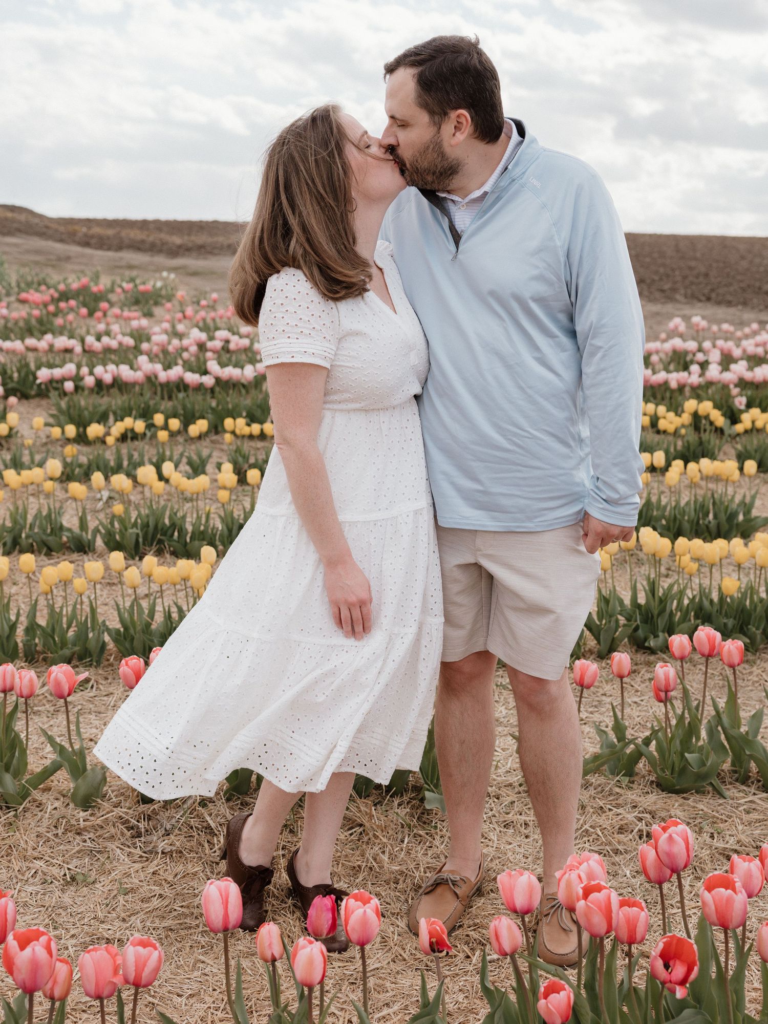 Couple sharing a kiss in a colorful tulip field with pink and yellow blooms on a cloudy day.