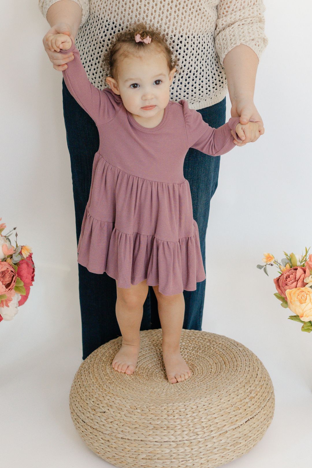 A small child in a pink dress stands on a woven mat with flowers in the background.