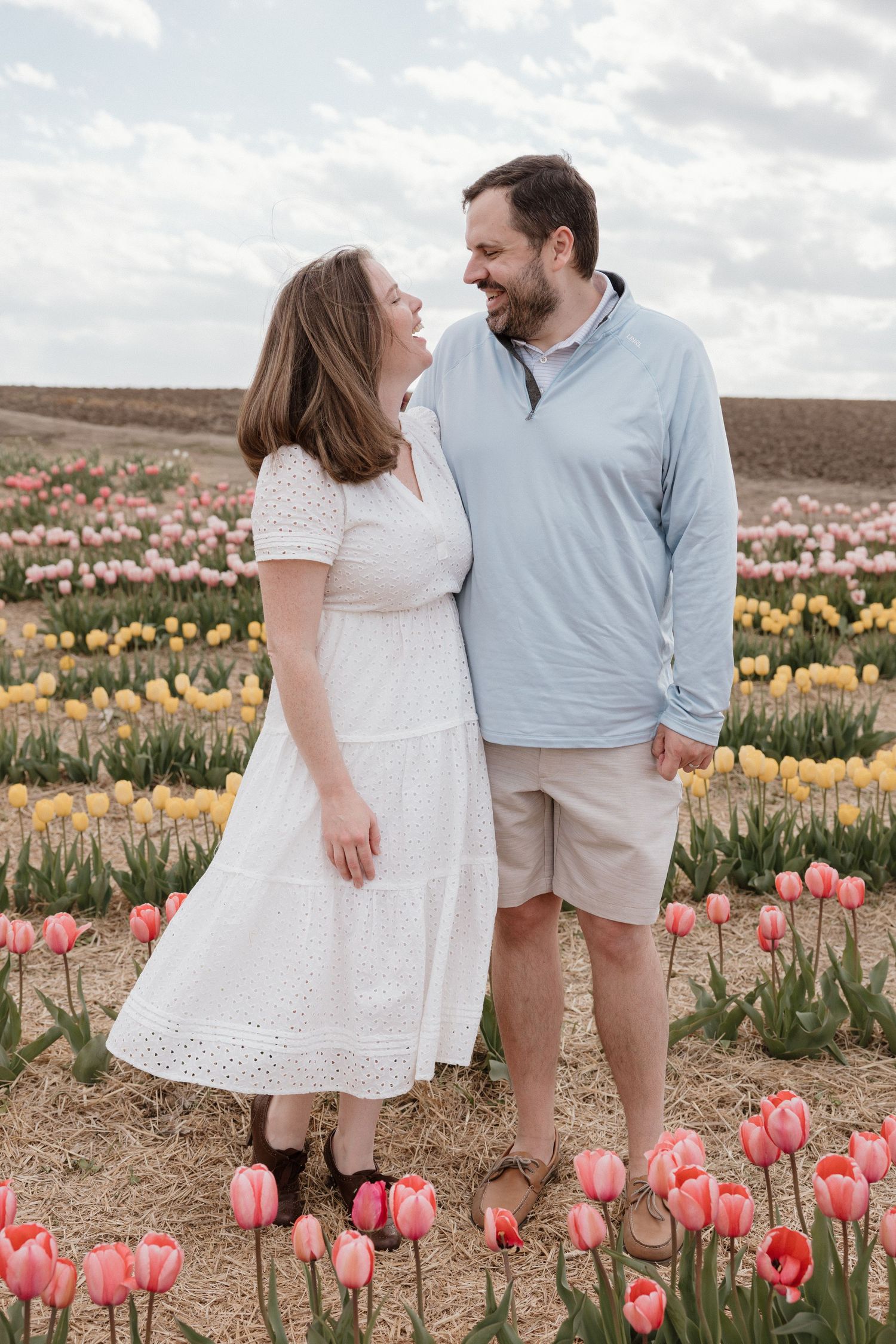Couple smiling at each other in a colorful tulip field with pink and yellow blooms under a cloudy sky.