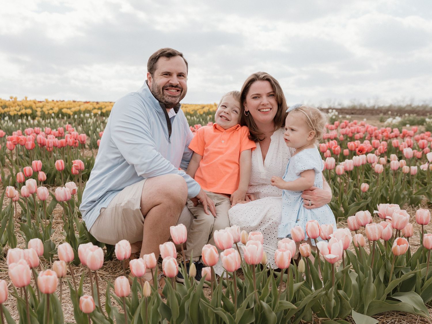 Family of four smiling among pink and yellow tulips in a blooming flower field on a cloudy spring day.