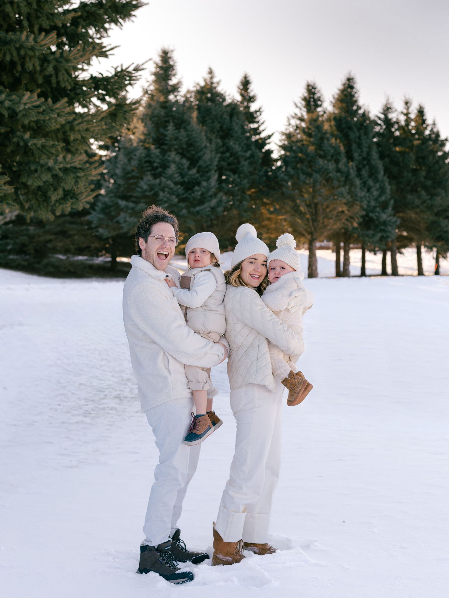 Family of four dressed in white poses joyfully in snowy landscape with evergreen trees in winter.