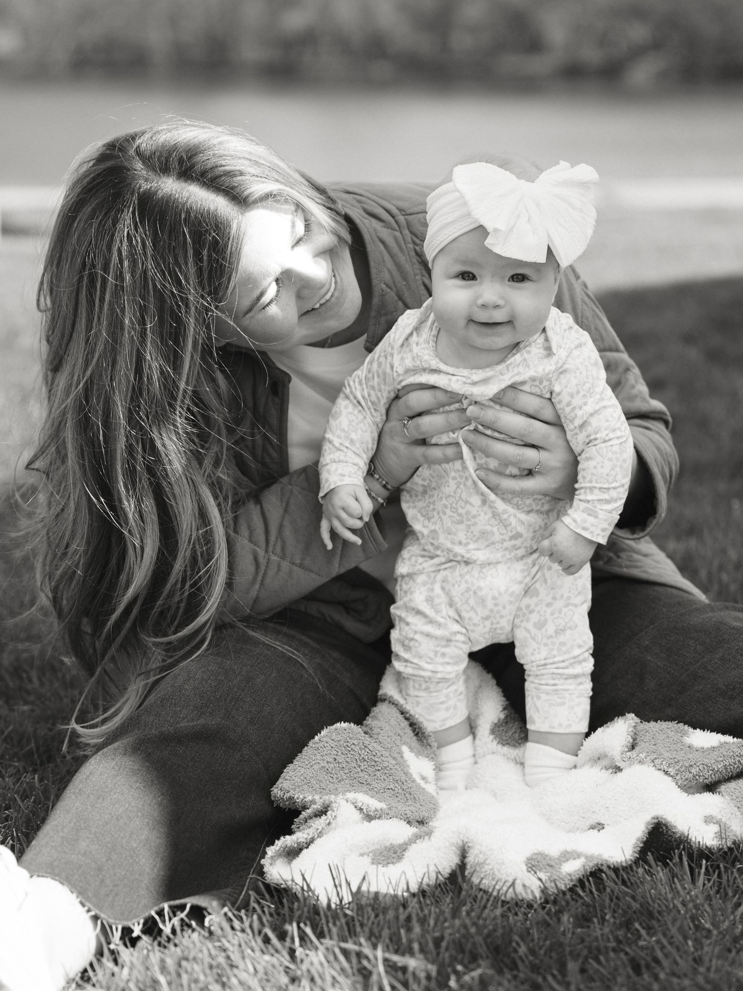 Black and white photo of a smiling woman holding a happy baby wearing a bow headband on a grassy lawn.