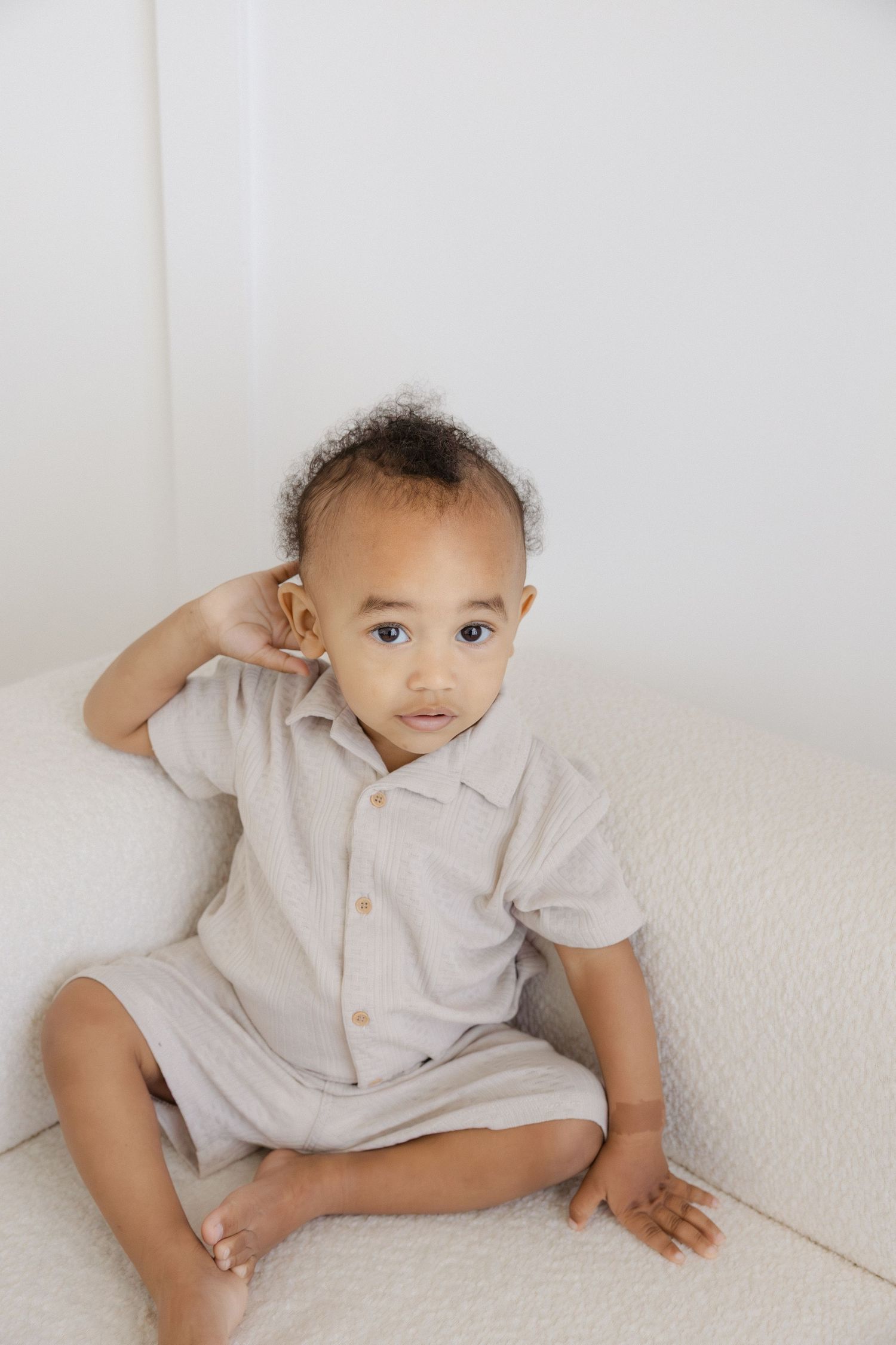 Adorable toddler in a beige linen romper sitting on a white couch, looking at the camera.