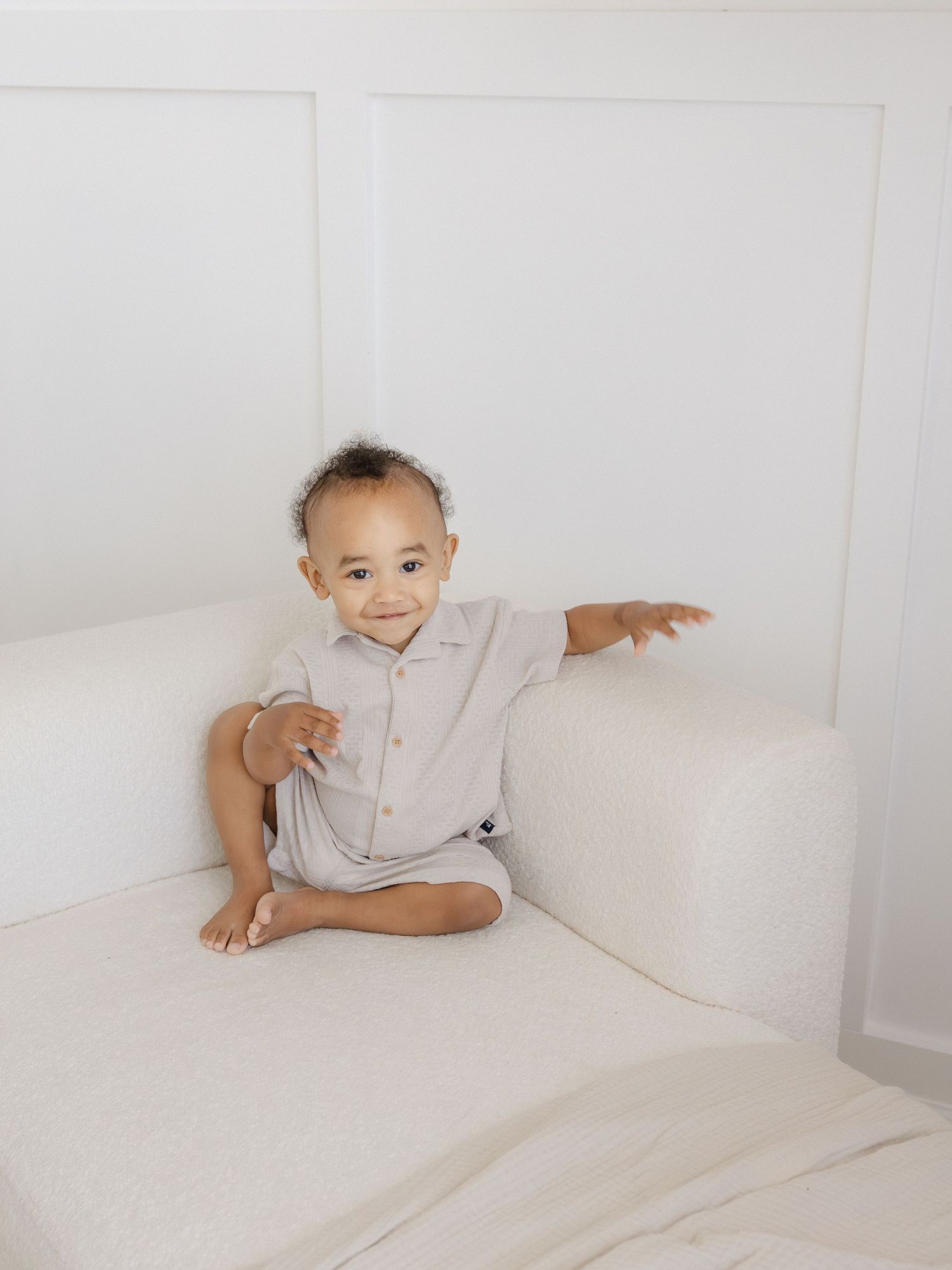Smiling baby in a light gray romper sitting cross-legged on a white couch in a bright, minimalist setting.