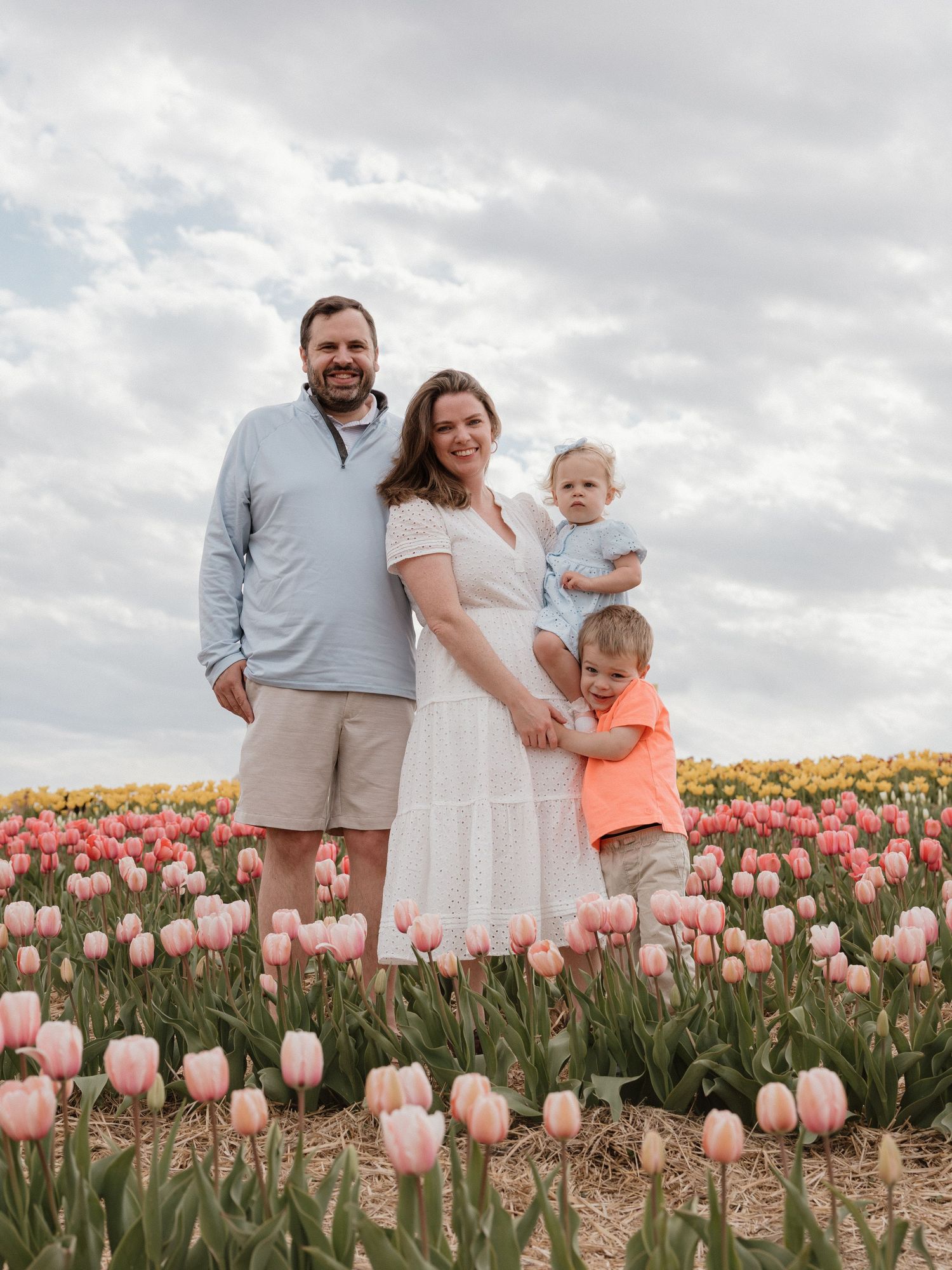 Family of four poses in a vibrant tulip field with pink and yellow blooms under a cloudy sky.