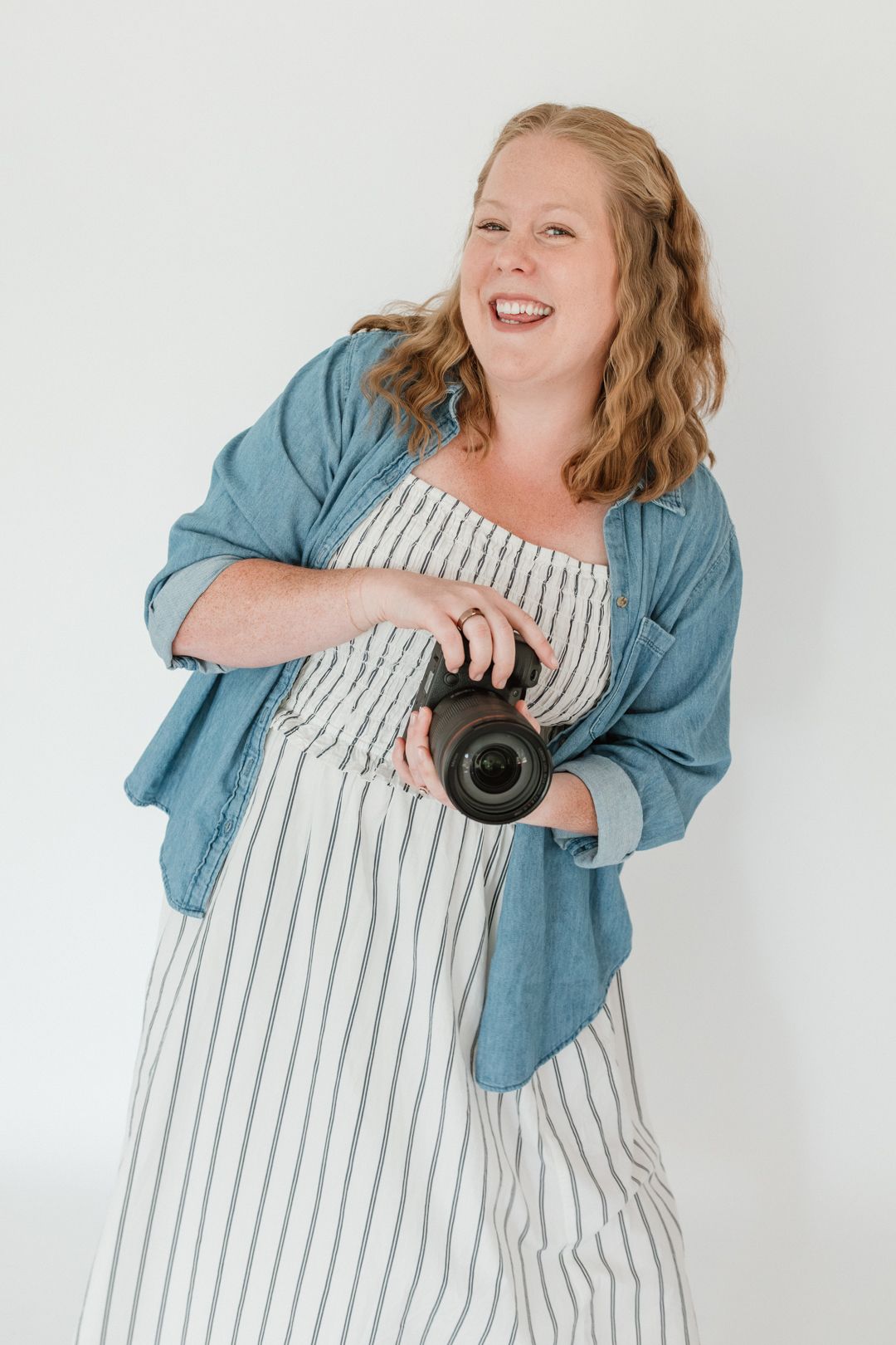 Smiling woman in striped dress and denim shirt holding a camera against a white background.