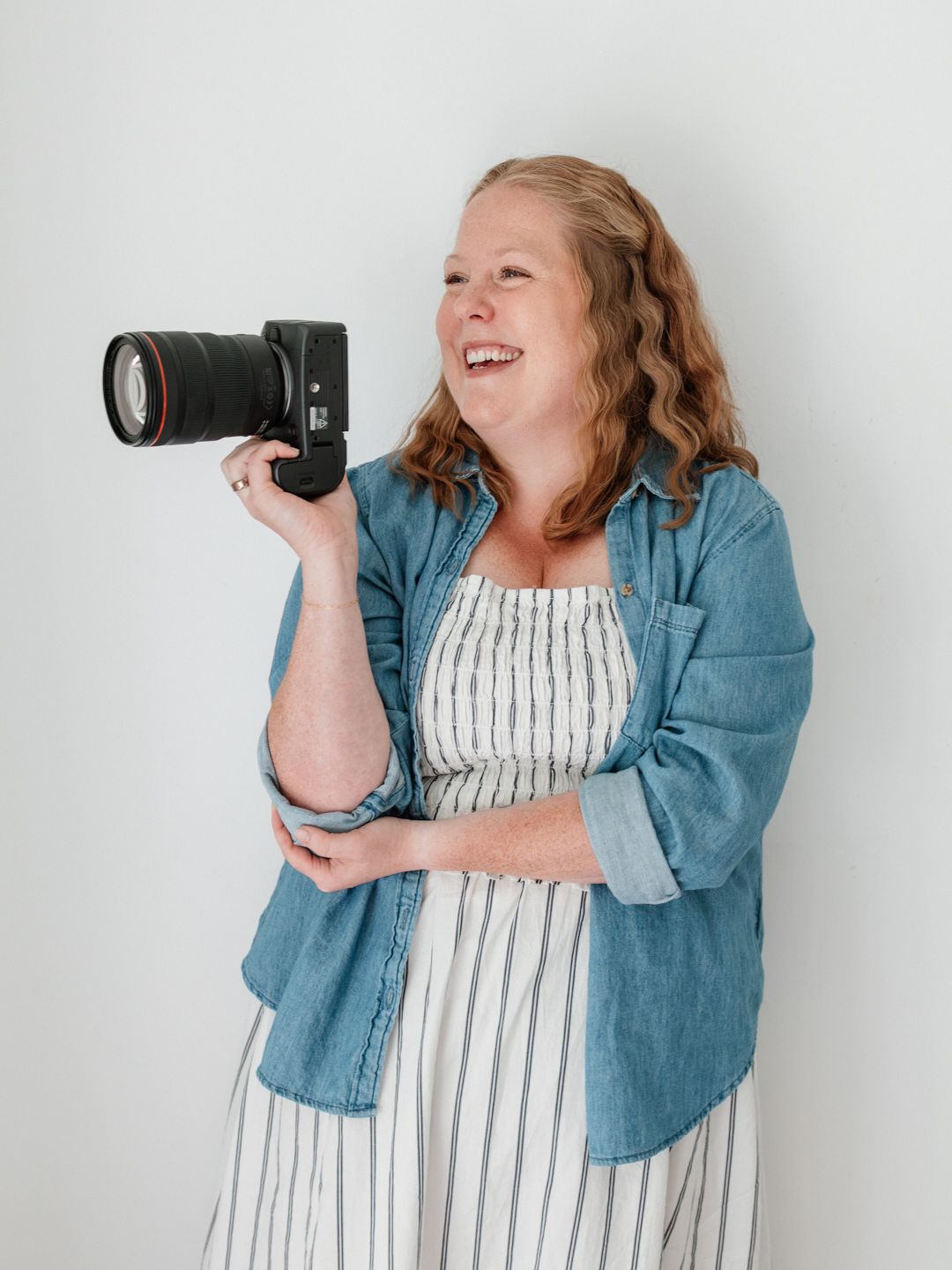 Smiling woman holding a DSLR camera wearing a denim shirt and striped dress against a white background.