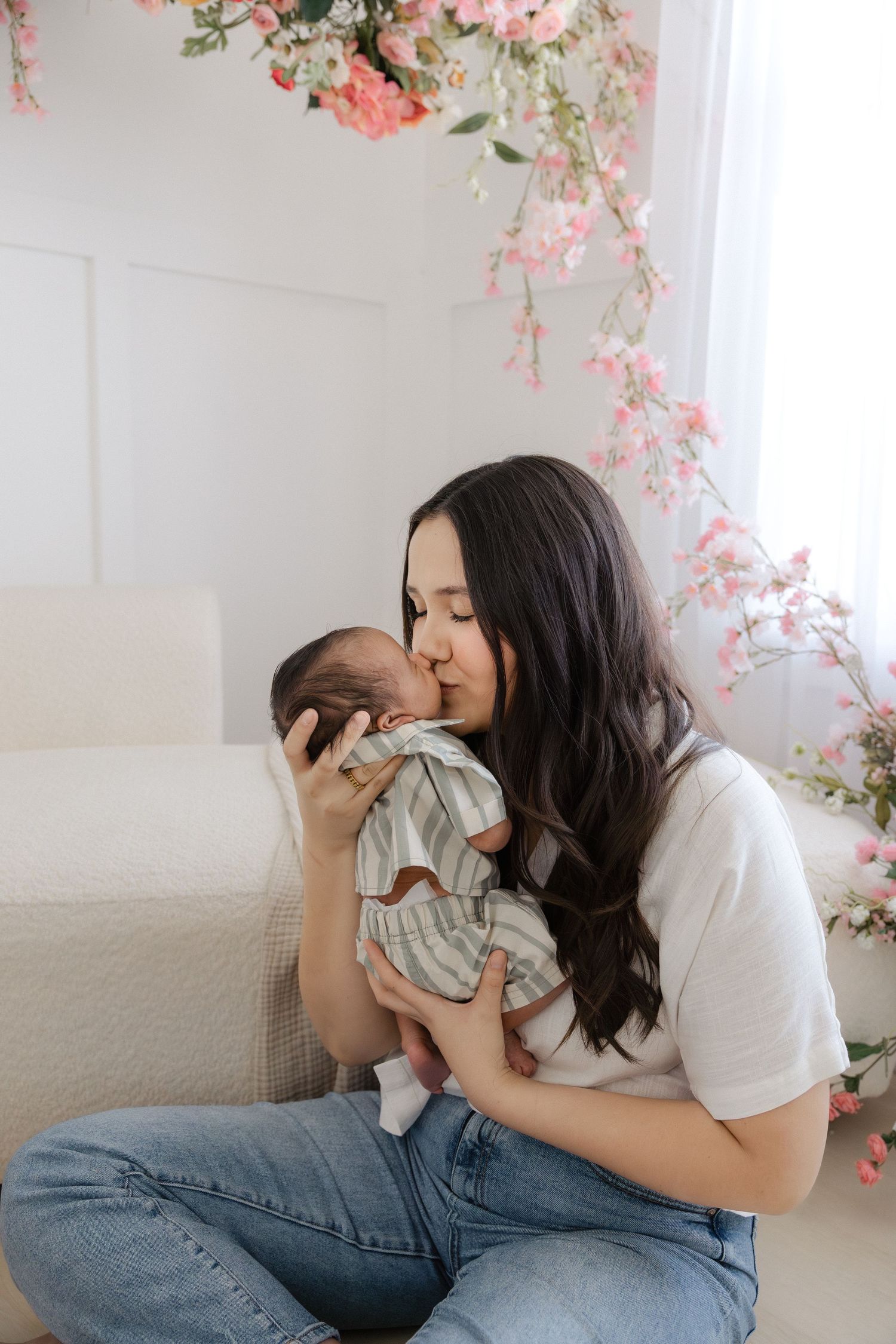 A mother gently kisses her newborn baby while sitting in a bright room adorned with pink floral decorations.