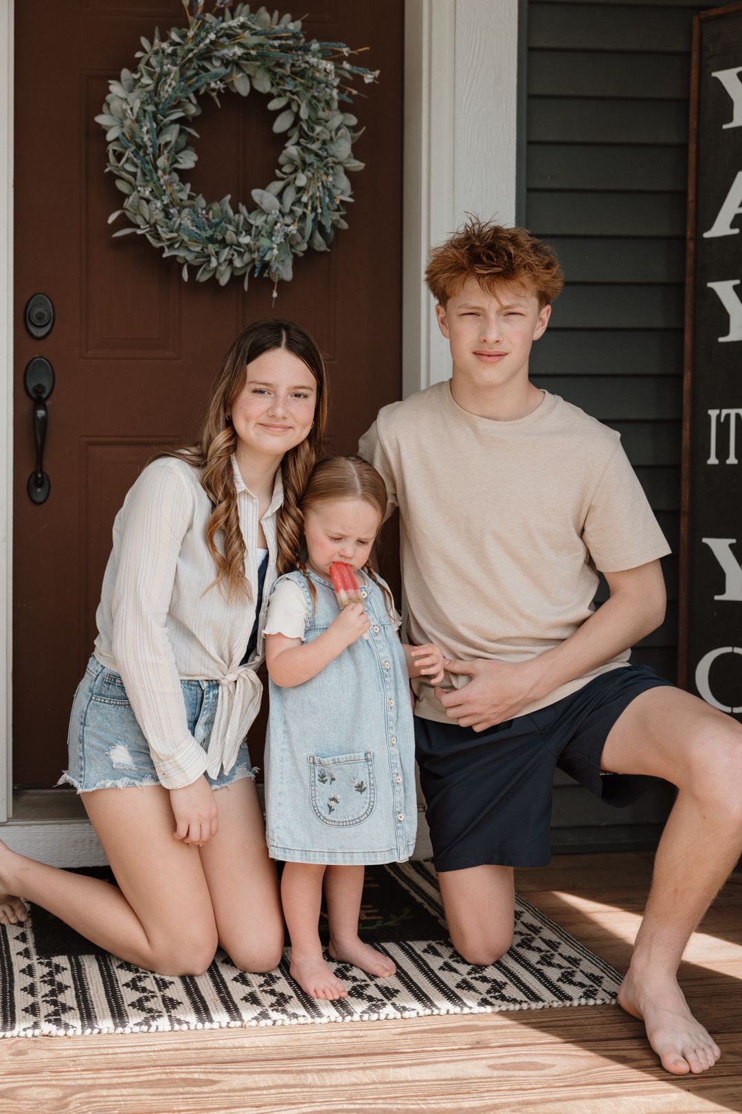 Teen girl and boy with young toddler sister sitting on front porch steps in casual summer outfits.