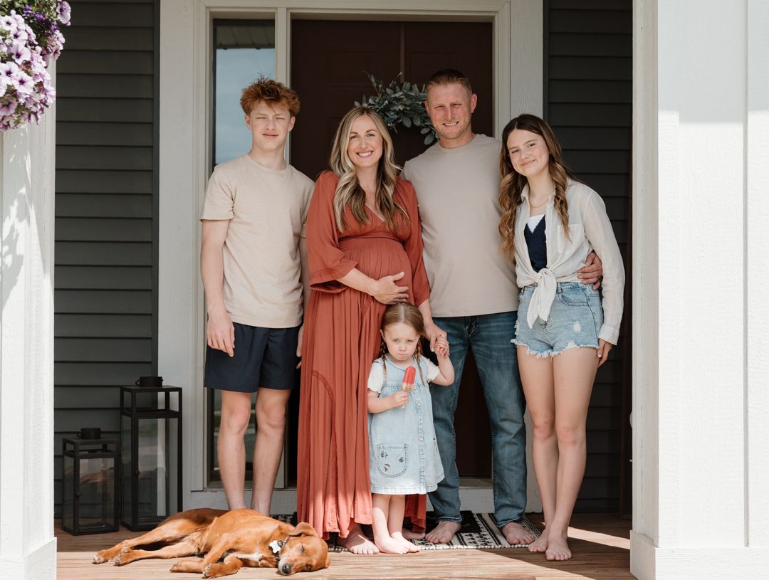 Family of five posing on front porch with dog, pregnant mother in rust dress, casual outdoor portrait.