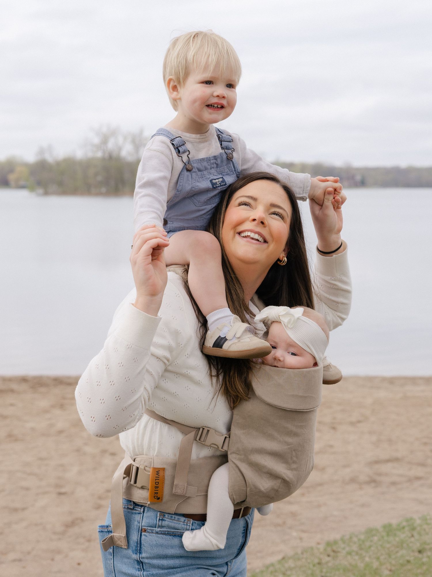 Smiling mom carries toddler on shoulders while babywearing infant in carrier near a lake outdoors.