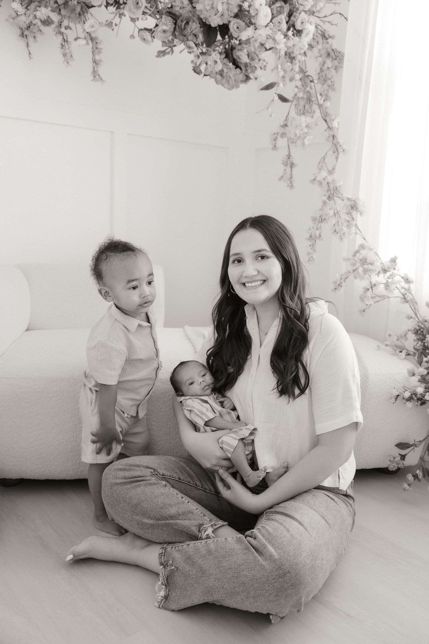 Black and white photo of a smiling mother sitting cross-legged holding a newborn, with a toddler standing beside her.