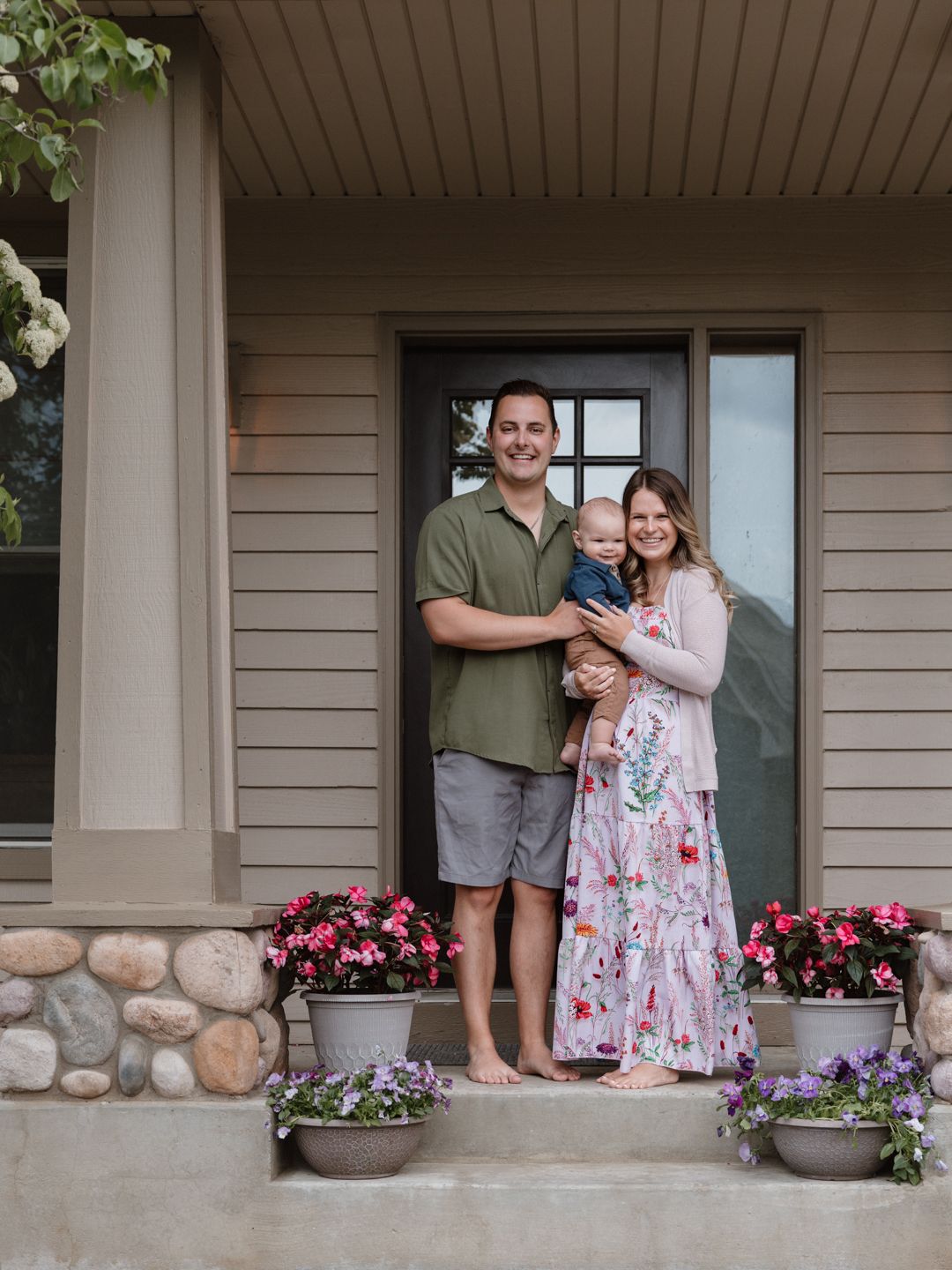 Happy young family with baby posing on front porch of suburban home with flower pots and stone accents.