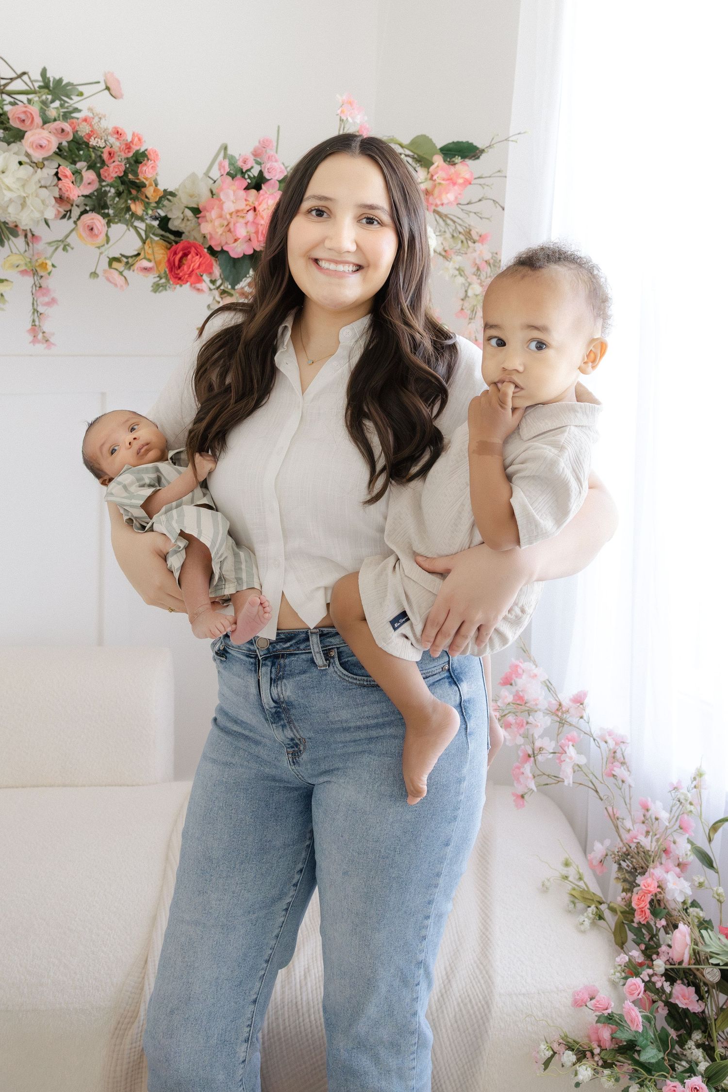 Smiling woman holding a newborn and a toddler in a bright floral studio setting, wearing jeans and a white top.