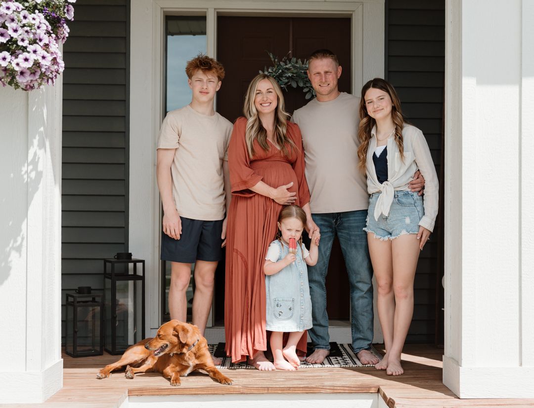 Family of five with pregnant mom in rust dress and golden dog pose on front porch steps.