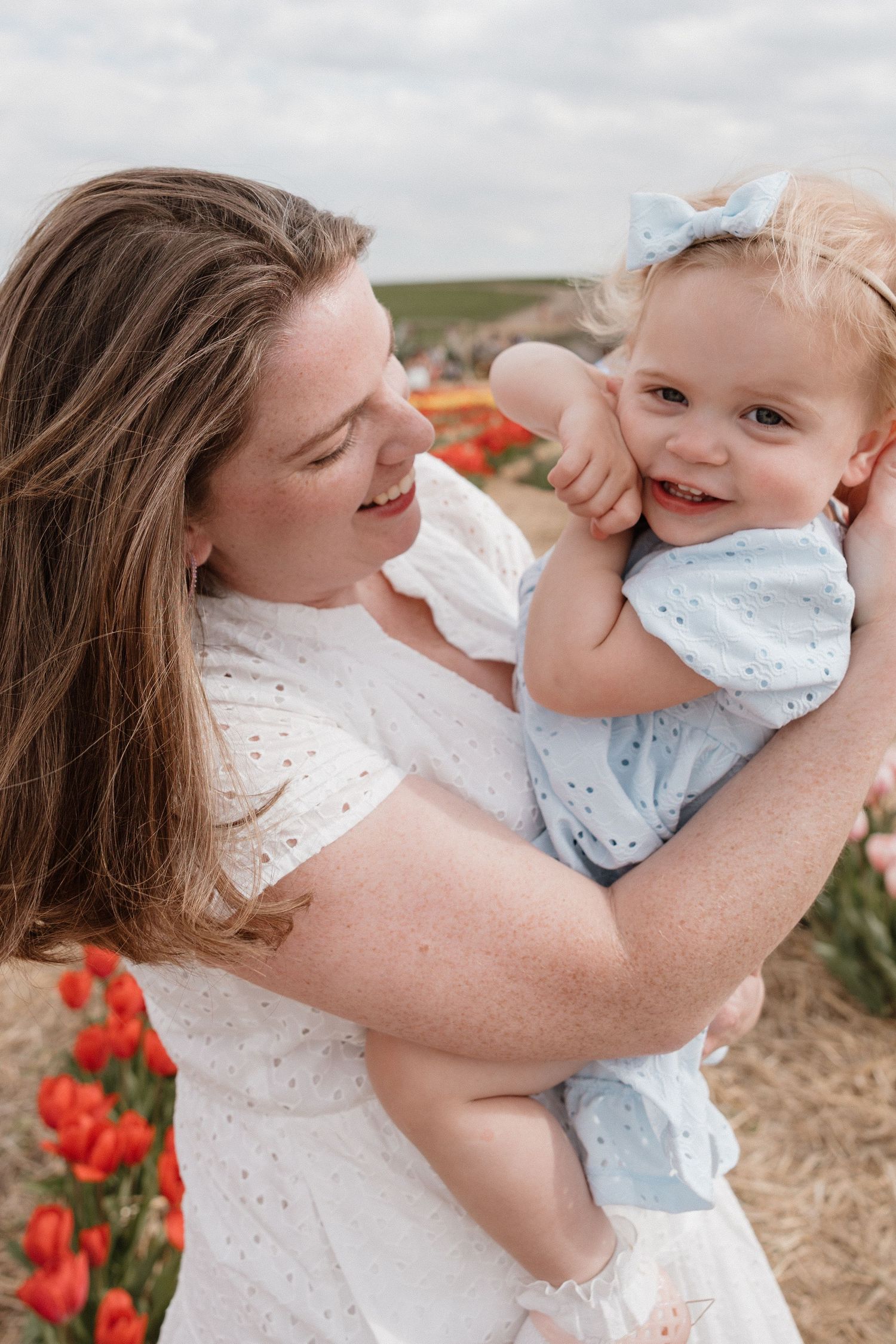 A smiling woman in a white dress holds a laughing toddler girl in a blue dress among red tulips.