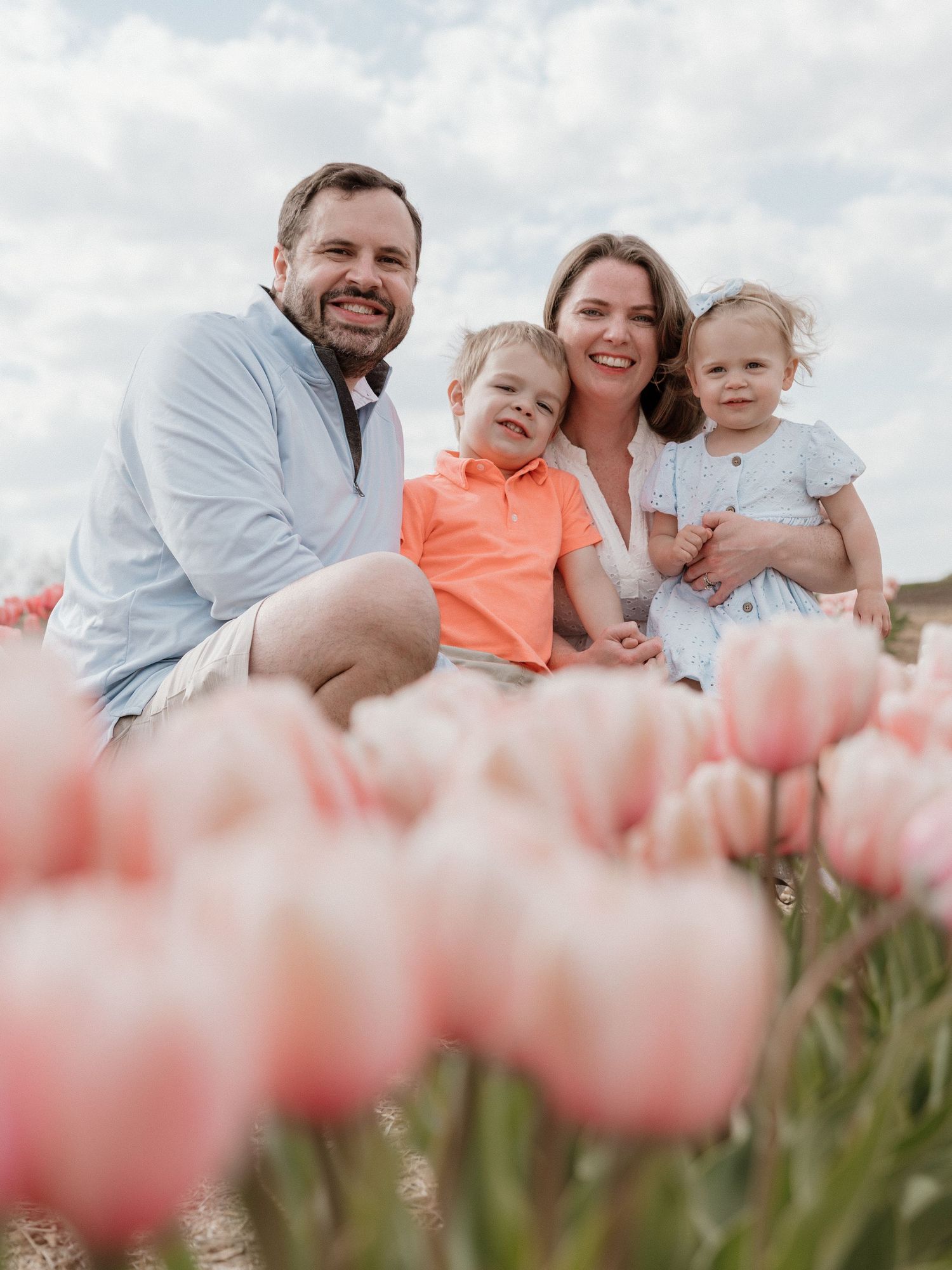 Family of four smiling in a tulip field, parents with young boy and toddler girl surrounded by pink blooms.