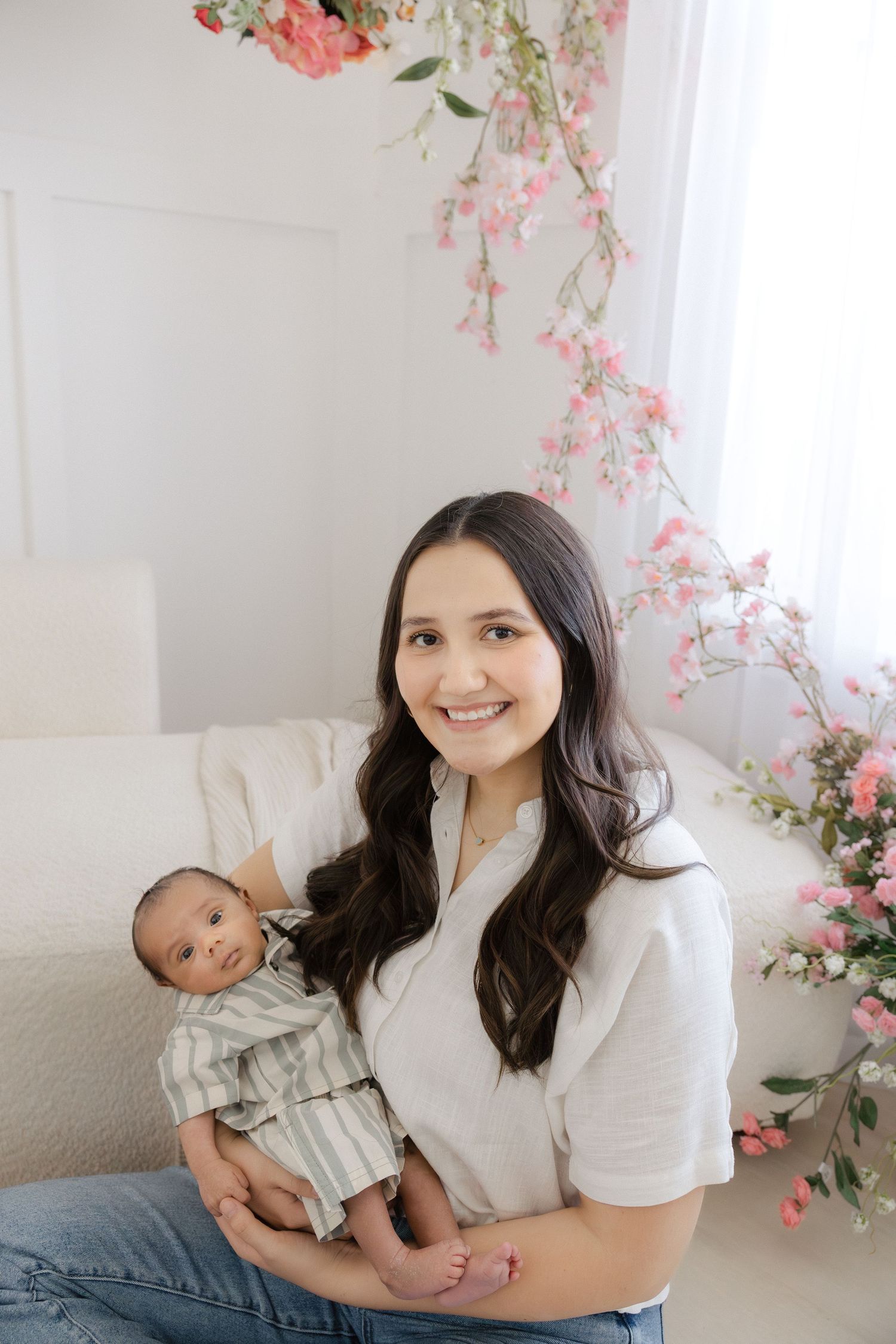 Smiling young mother holding her newborn baby in a bright white studio with pink floral decorations.