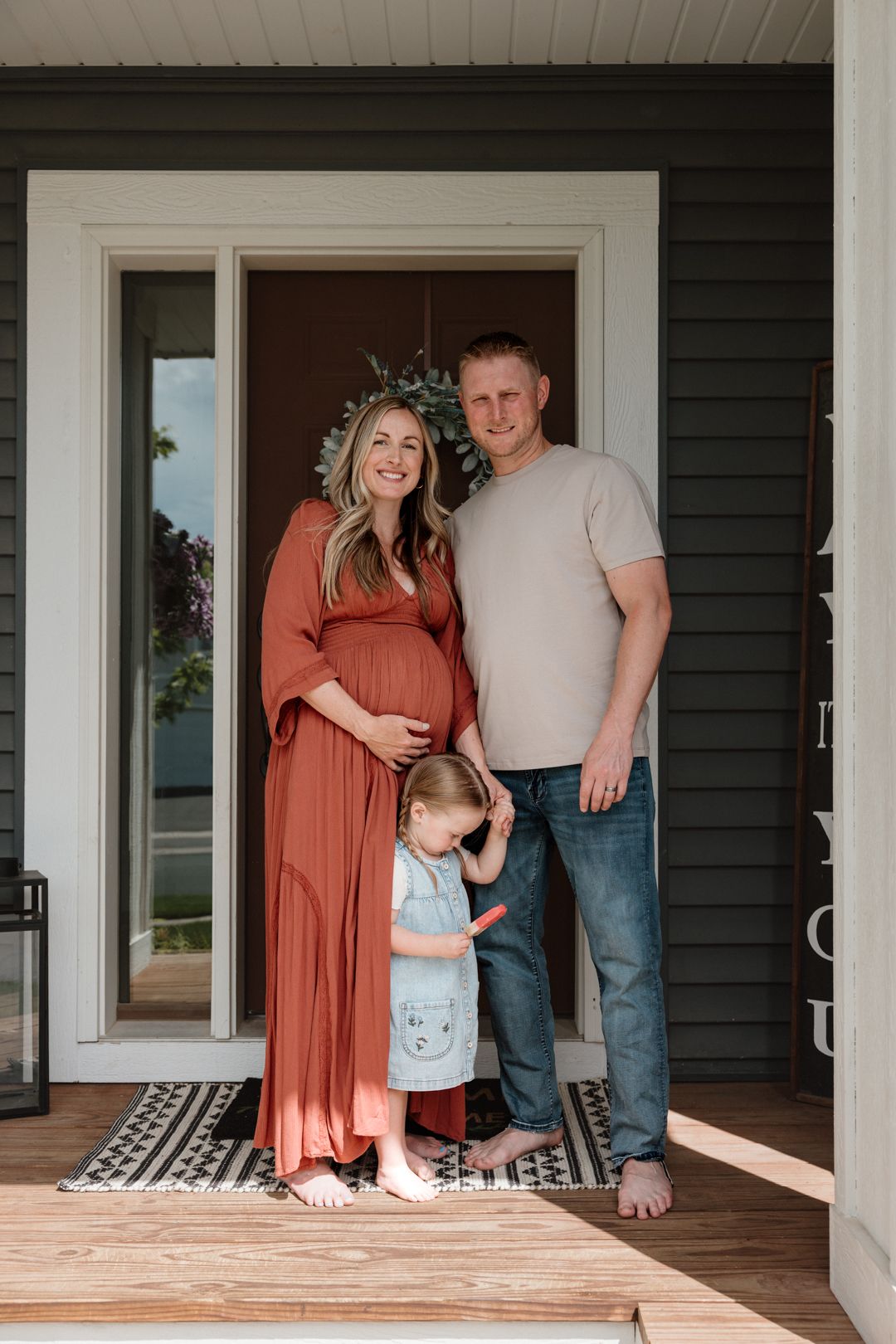 Pregnant woman in rust dress with husband and toddler girl posing on front porch for maternity photo.