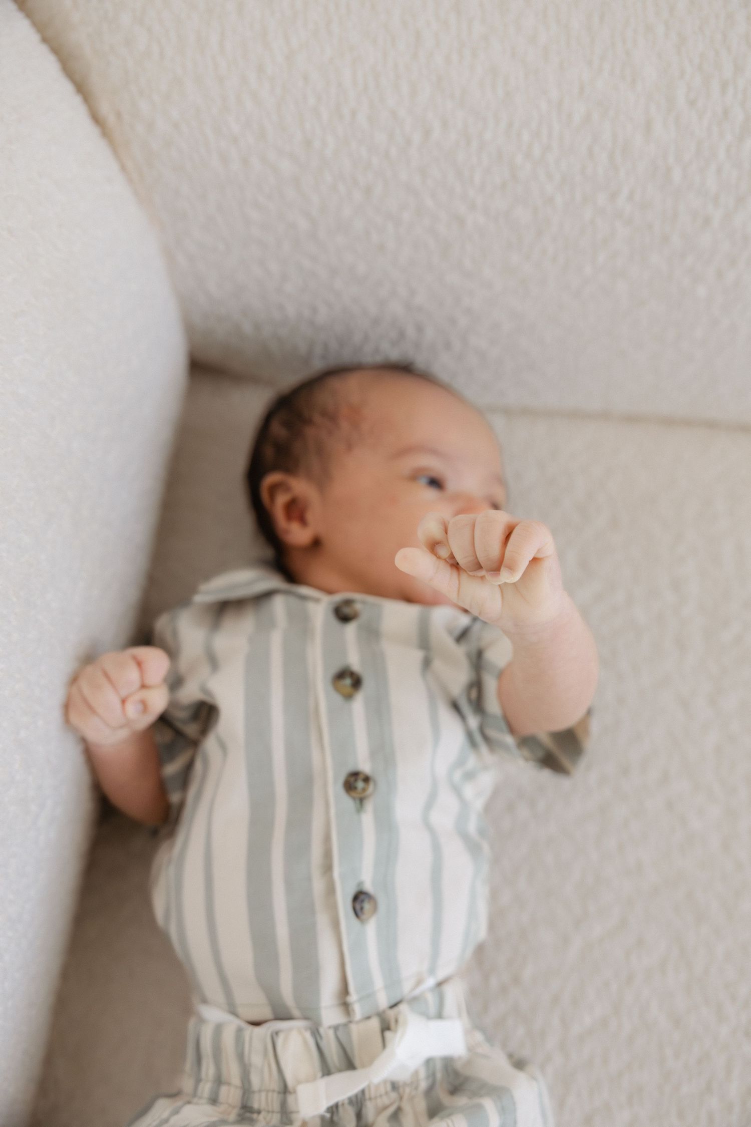 Newborn baby in striped sage green romper lying on cream sofa, hand raised to mouth.