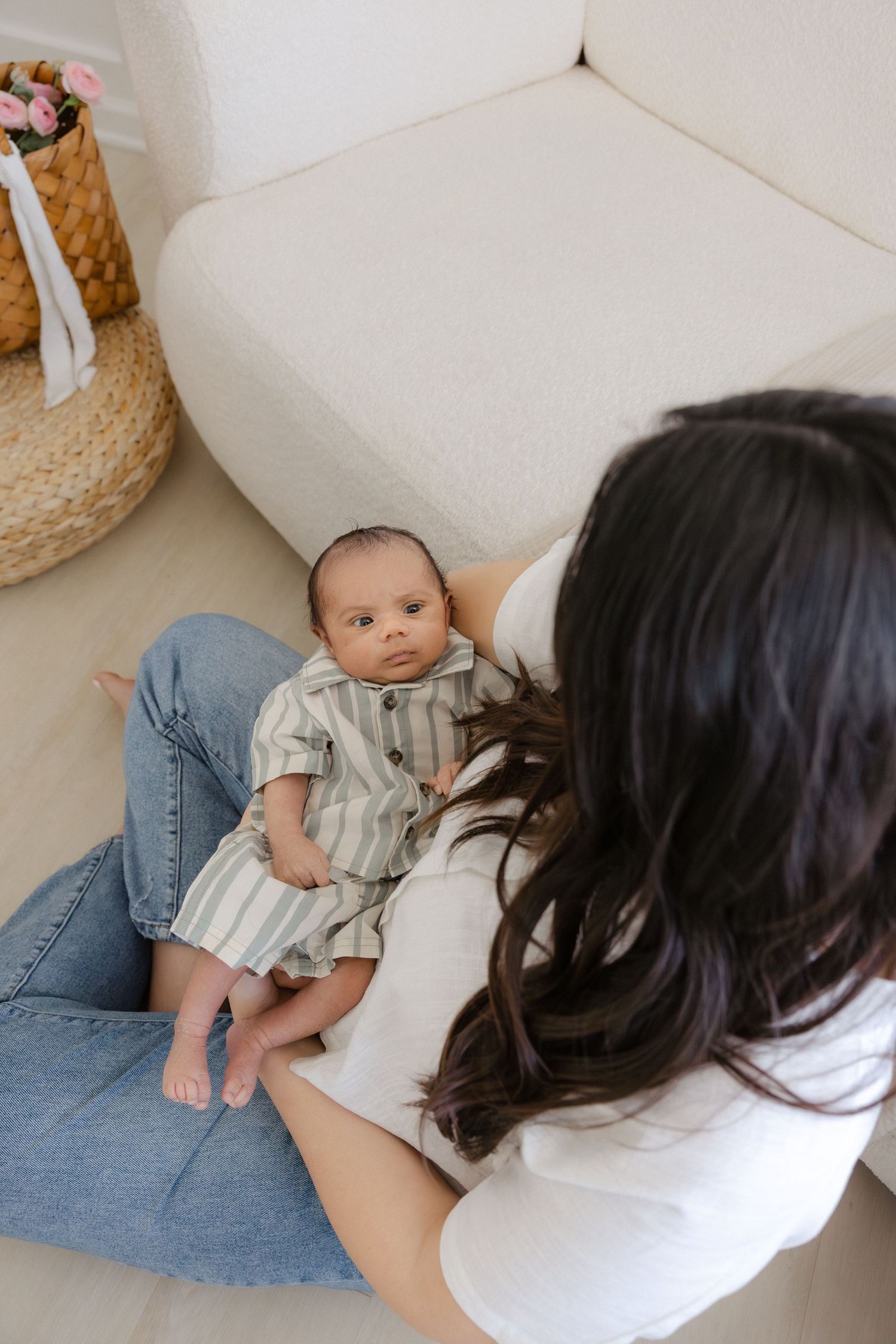 Mother sitting on floor cradling newborn baby in striped onesie against a white couch in a bright room.