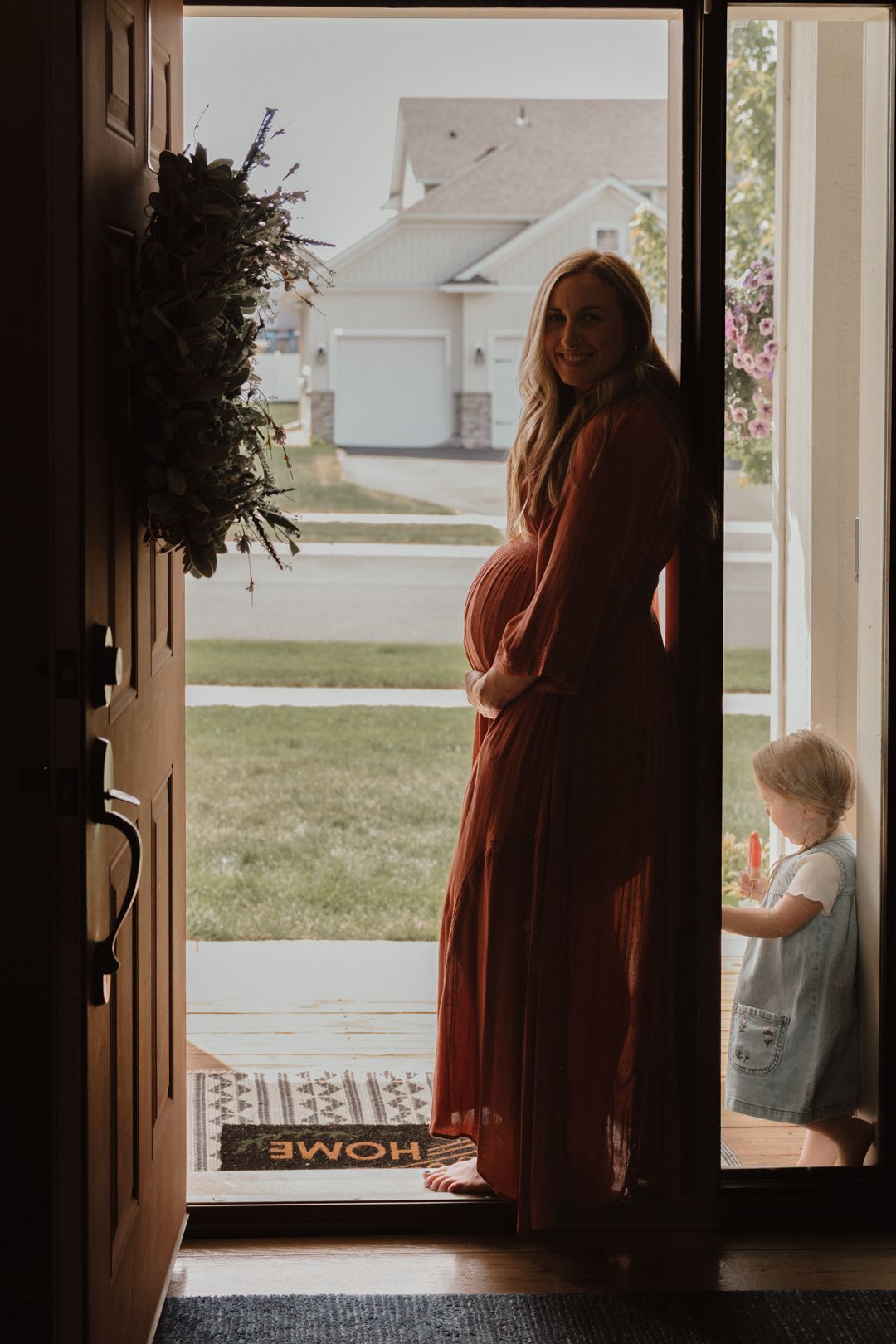 Pregnant woman in rust dress stands in open doorway smiling, with toddler beside her and a wreath on the door.
