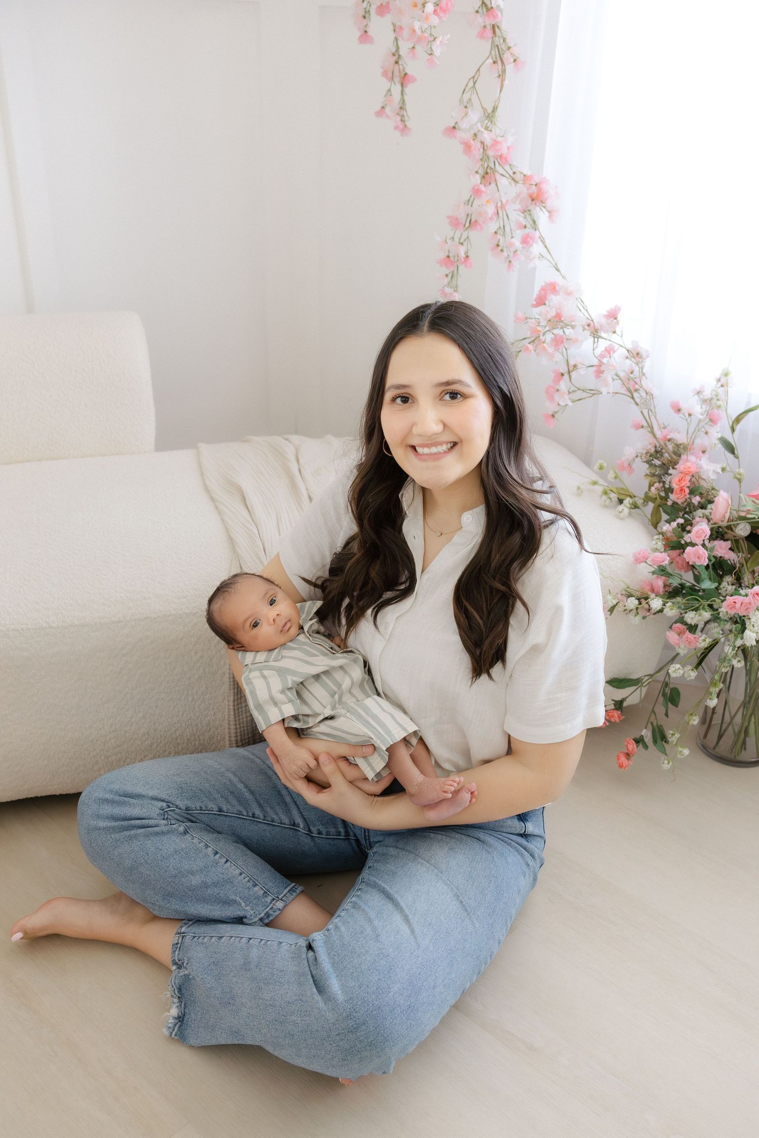 Young mother sitting on floor holding newborn baby, smiling, surrounded by pink flowers in bright white room.