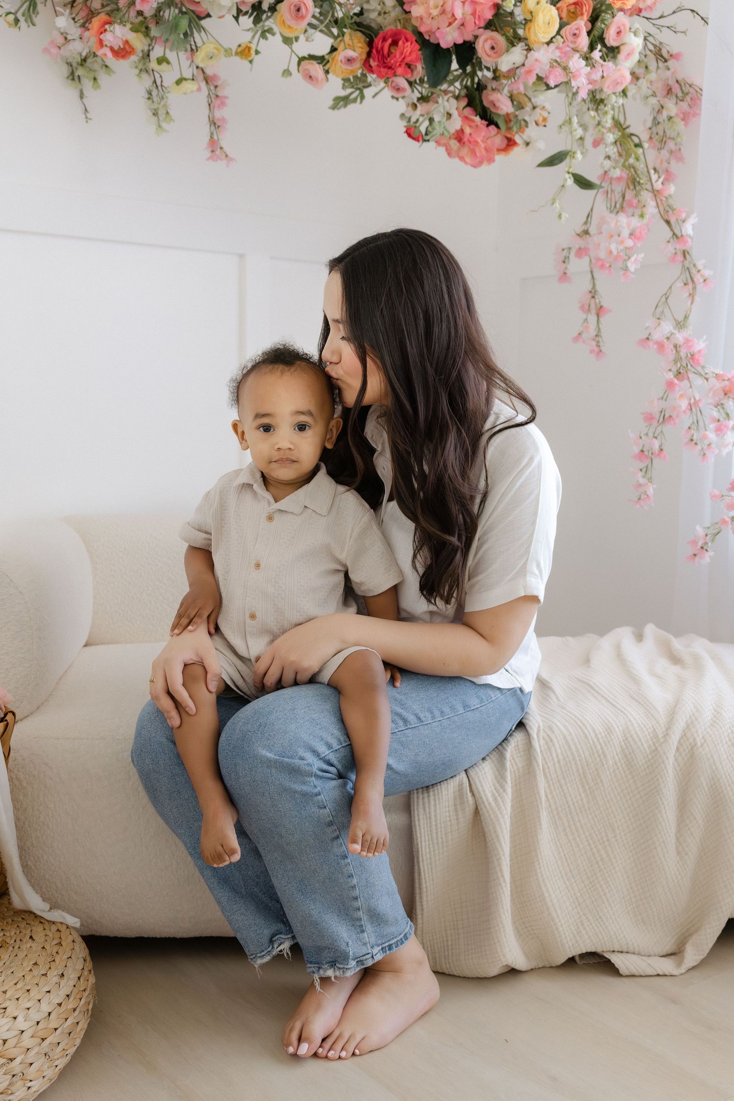 A mother kisses her toddler son on the head while sitting on a cream sofa beneath a floral arrangement.