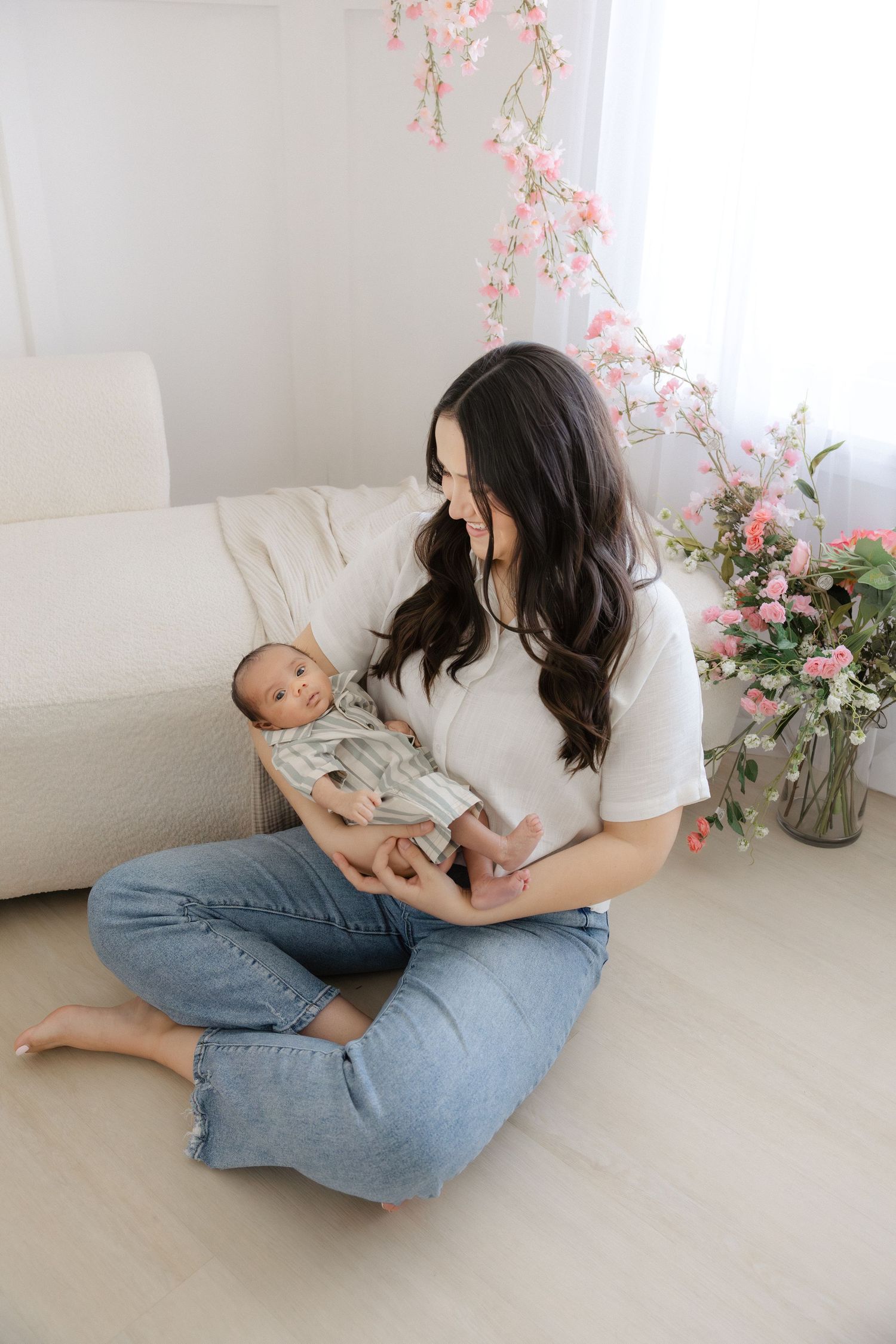 A young mother with dark hair sits cross-legged on a light wood floor, lovingly cradling her newborn baby.
