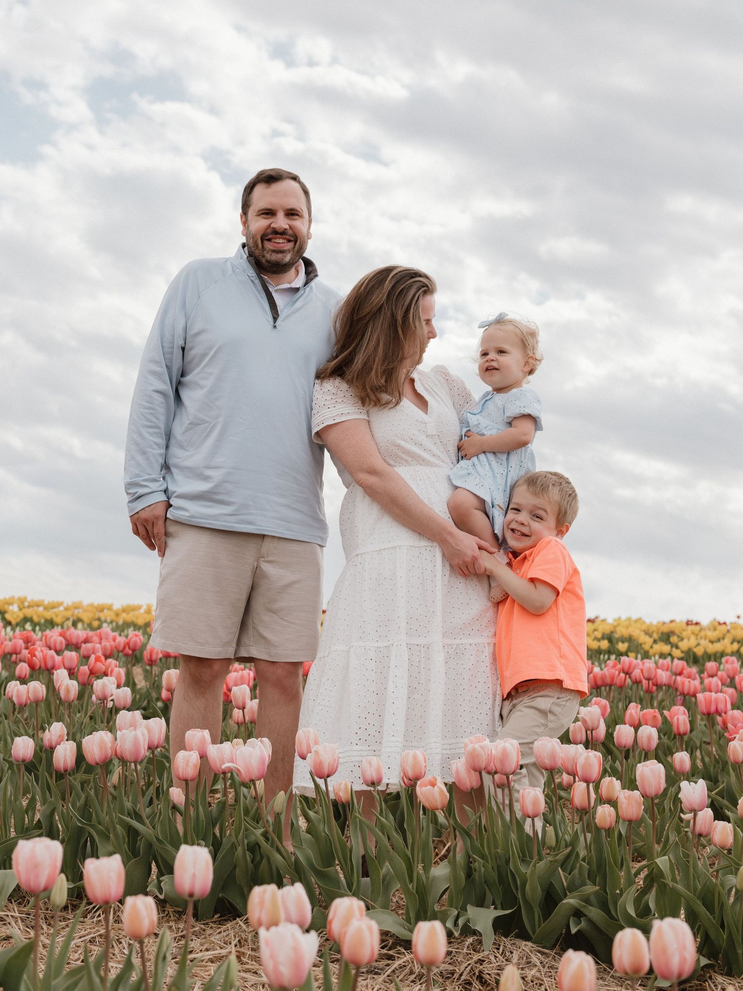 Family of four posing in a vibrant tulip field with pink and yellow blooms under a cloudy spring sky.