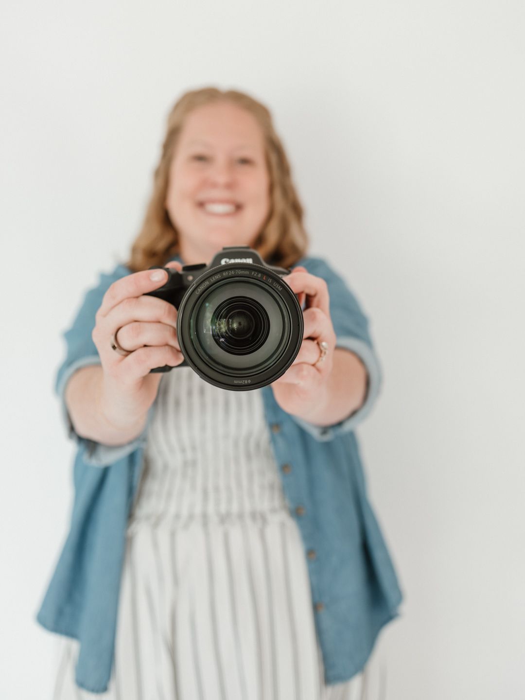 Smiling woman holding a Canon DSLR camera toward the viewer against a white background.