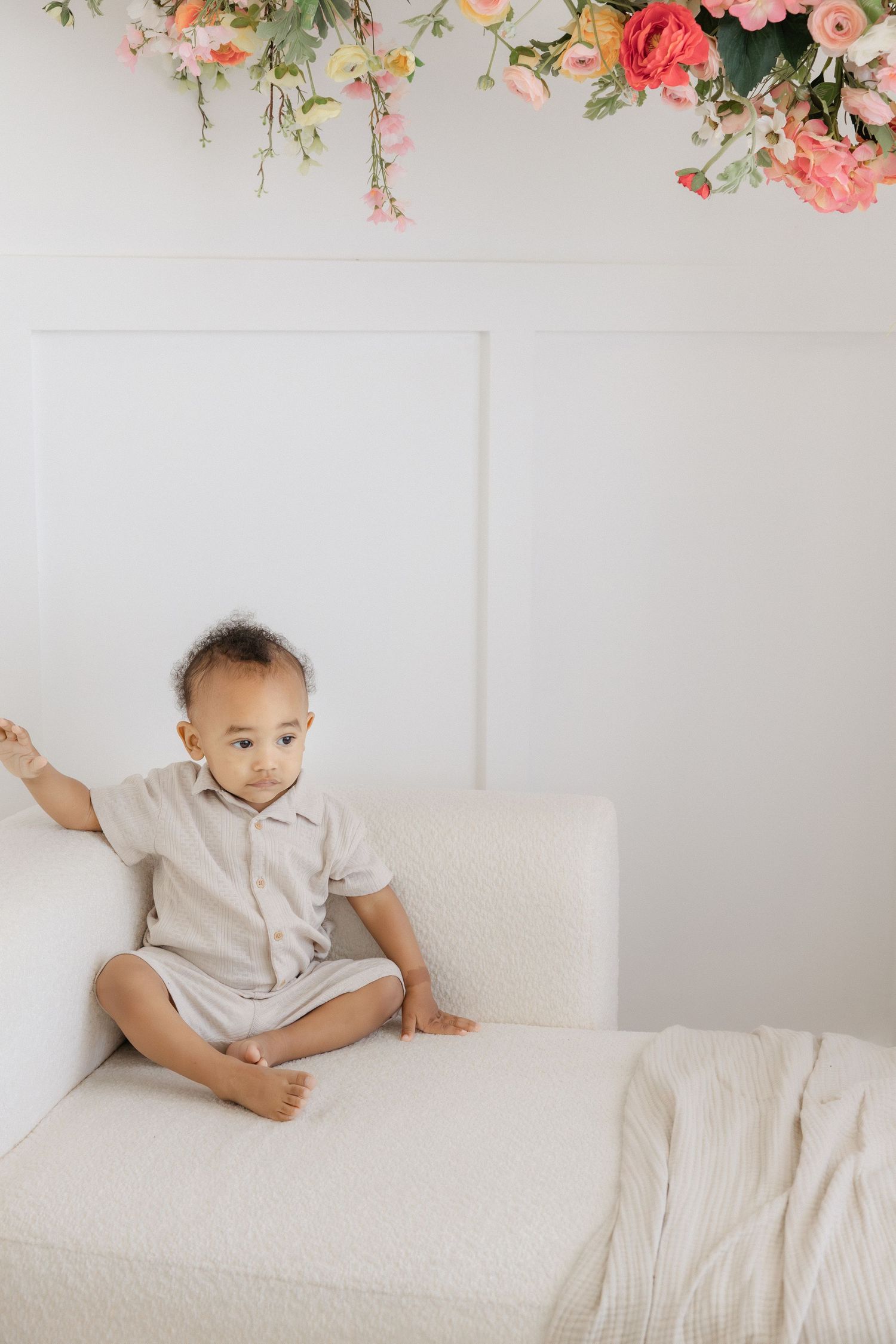 A toddler in a beige linen outfit sits cross-legged on a white sofa beneath pink floral decorations.
