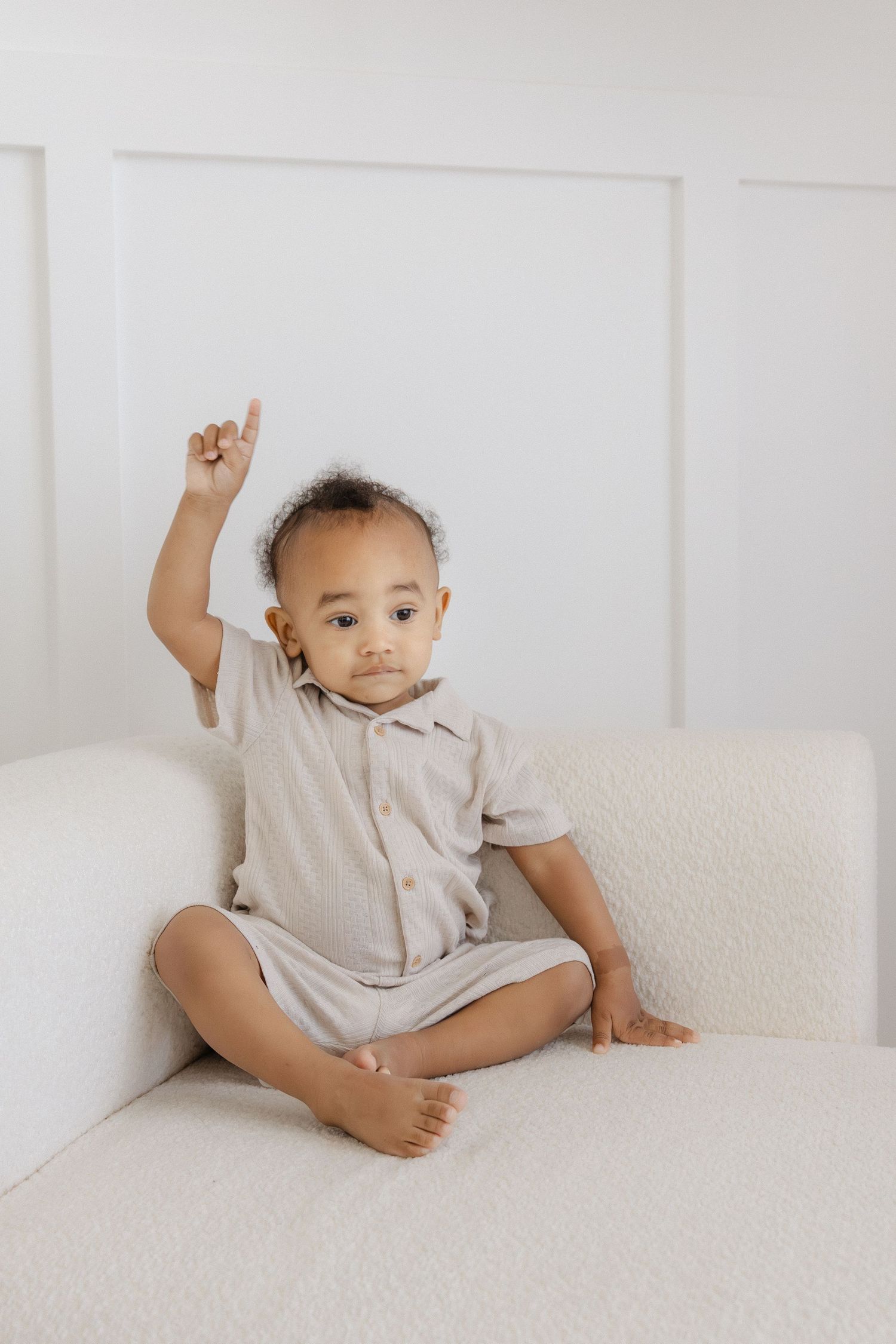 Cute toddler in beige romper sitting cross-legged on white sofa with one finger raised up.