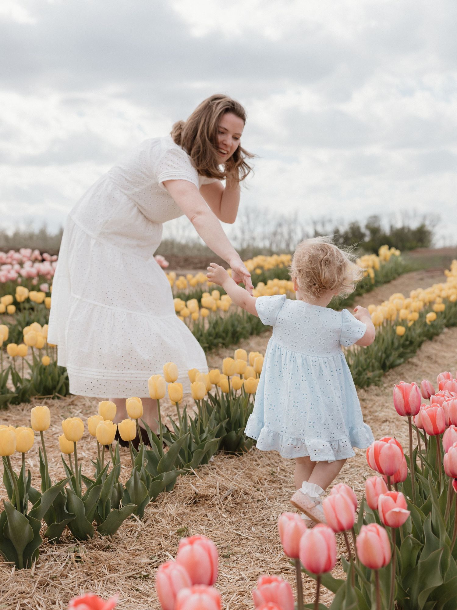 A mother in a white dress plays with her toddler in a colorful tulip field on a cloudy spring day.