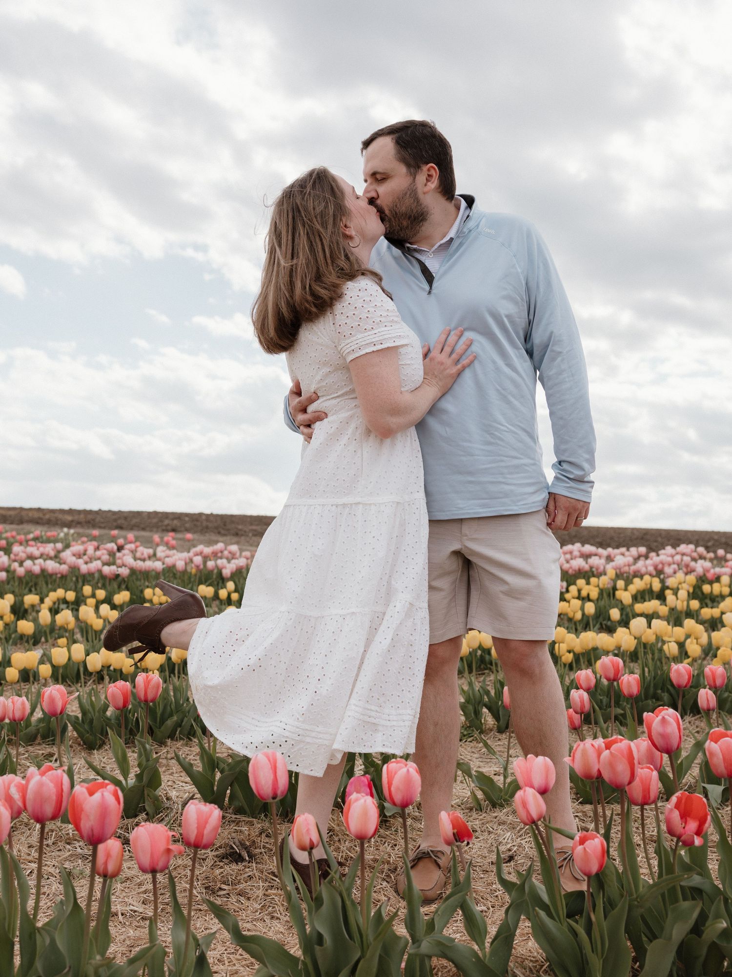 Couple kissing in a colorful tulip field with pink and yellow blooms under a cloudy sky.
