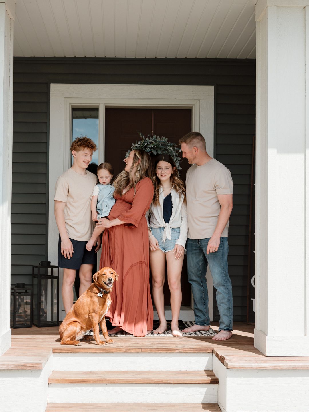 Family of five with a golden retriever posing on a porch, mother in rust dress wearing floral crown.