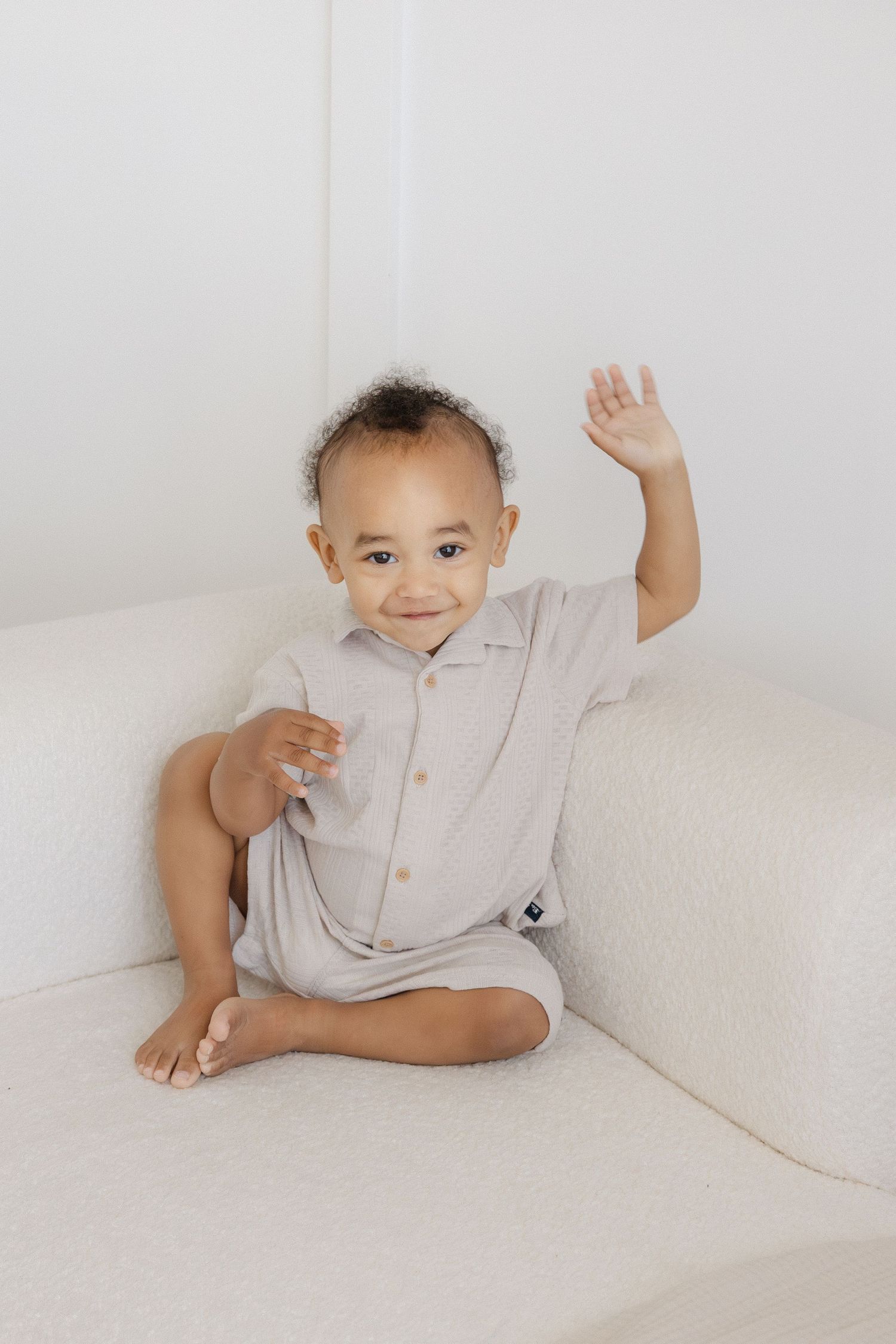 Smiling baby in a gray romper sits on a white couch, waving one hand up in the air.