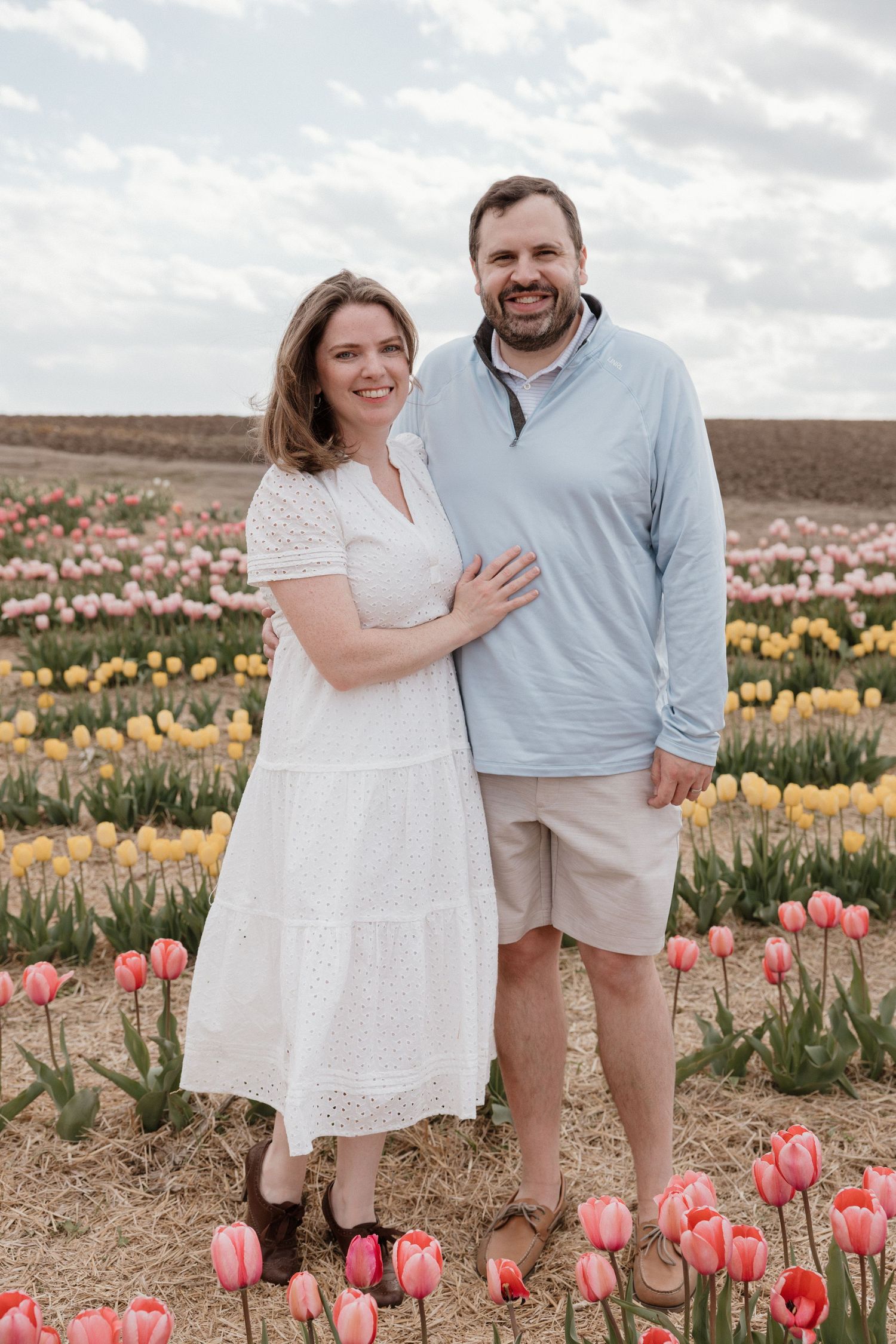 Couple smiling together in a colorful tulip field with pink and yellow flowers under a cloudy sky.