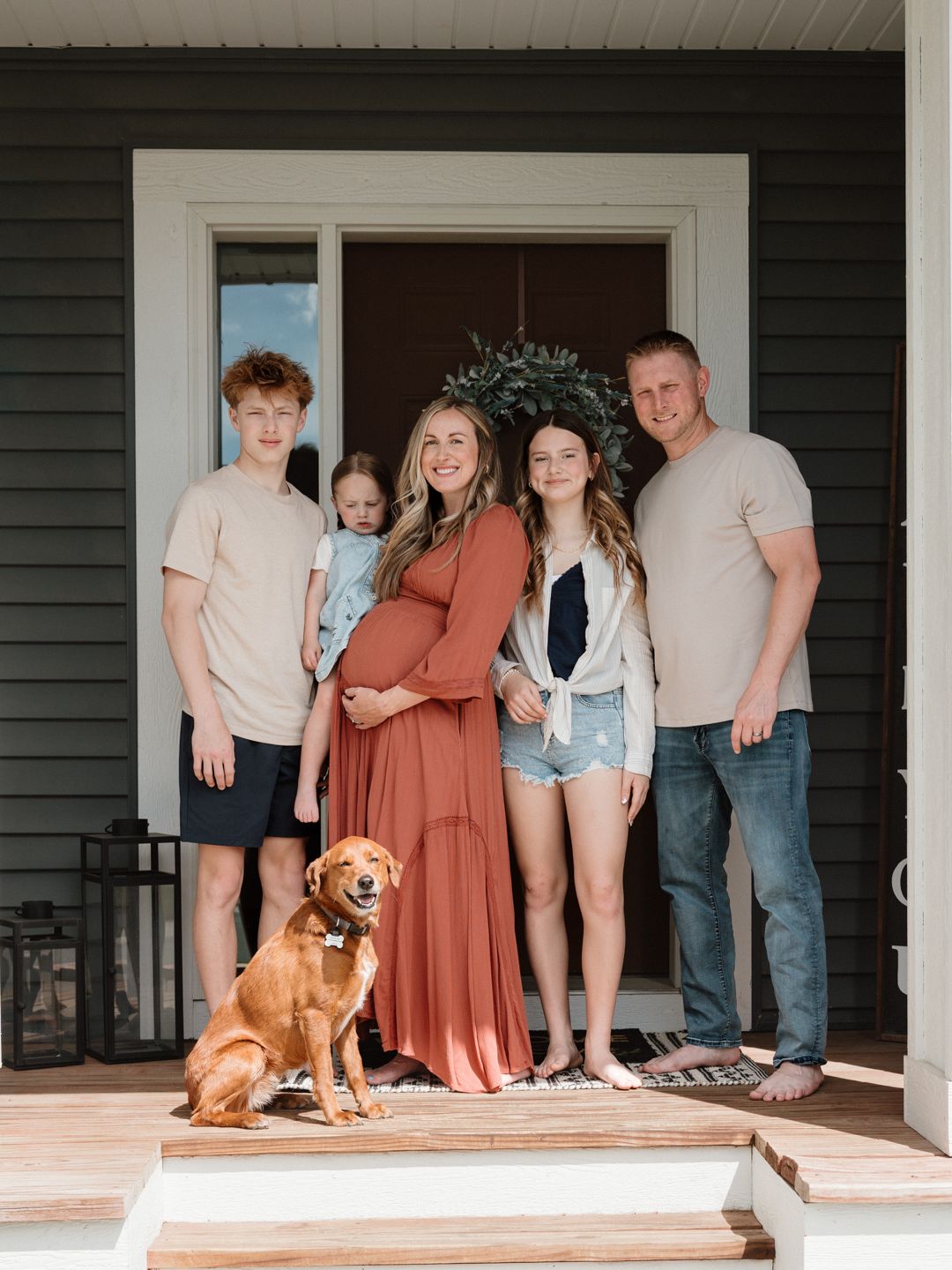Pregnant woman poses with family and golden retriever dog on home front porch for maternity photo session.