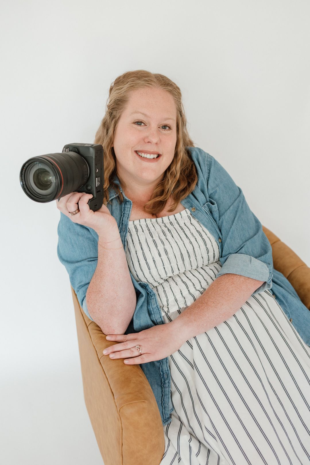 Smiling woman holding a DSLR camera, wearing a striped dress and denim shirt, seated on a tan chair against a white background.