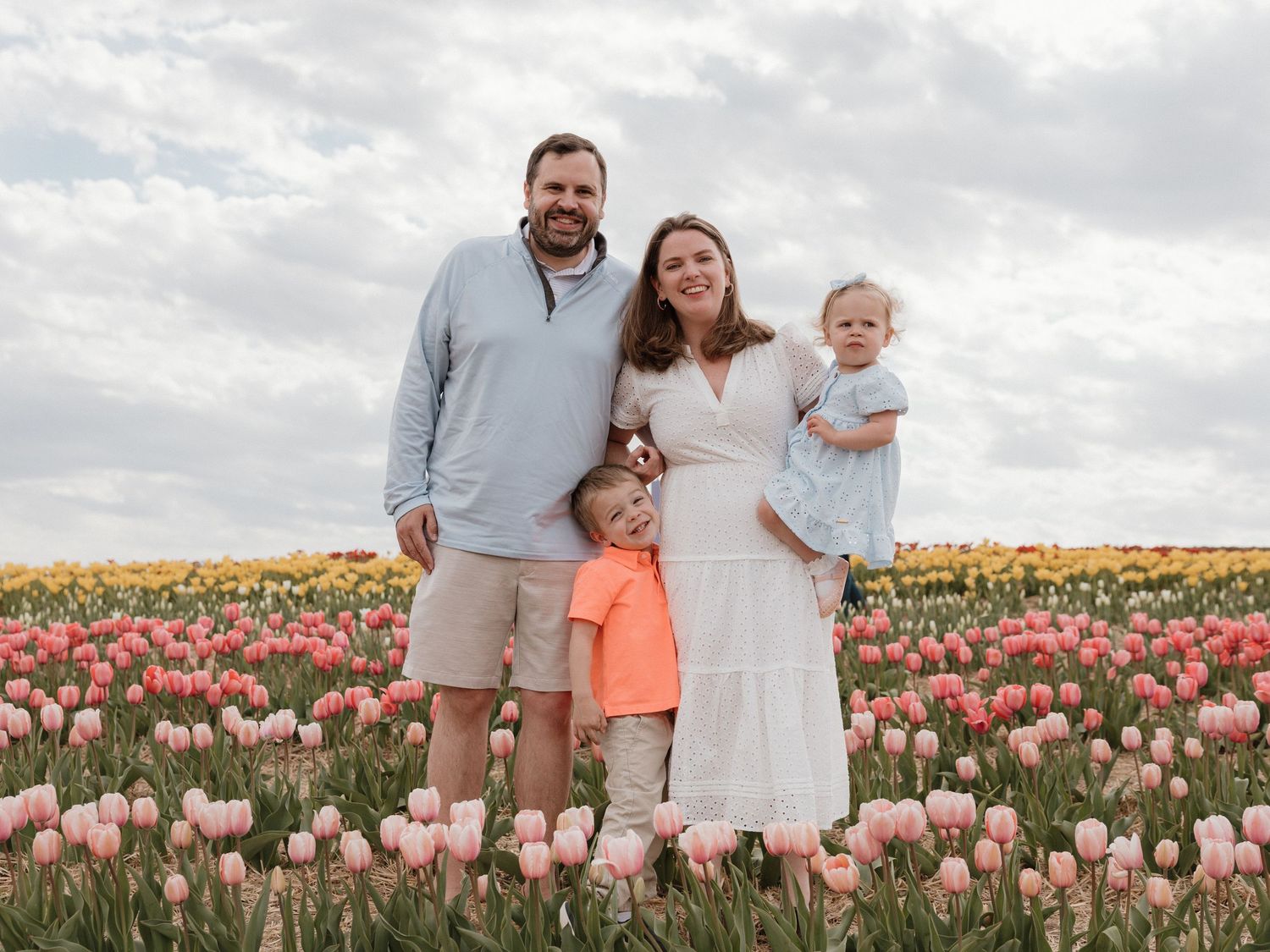 Family of four posing in a vibrant tulip field with pink and yellow blooms under a cloudy sky.